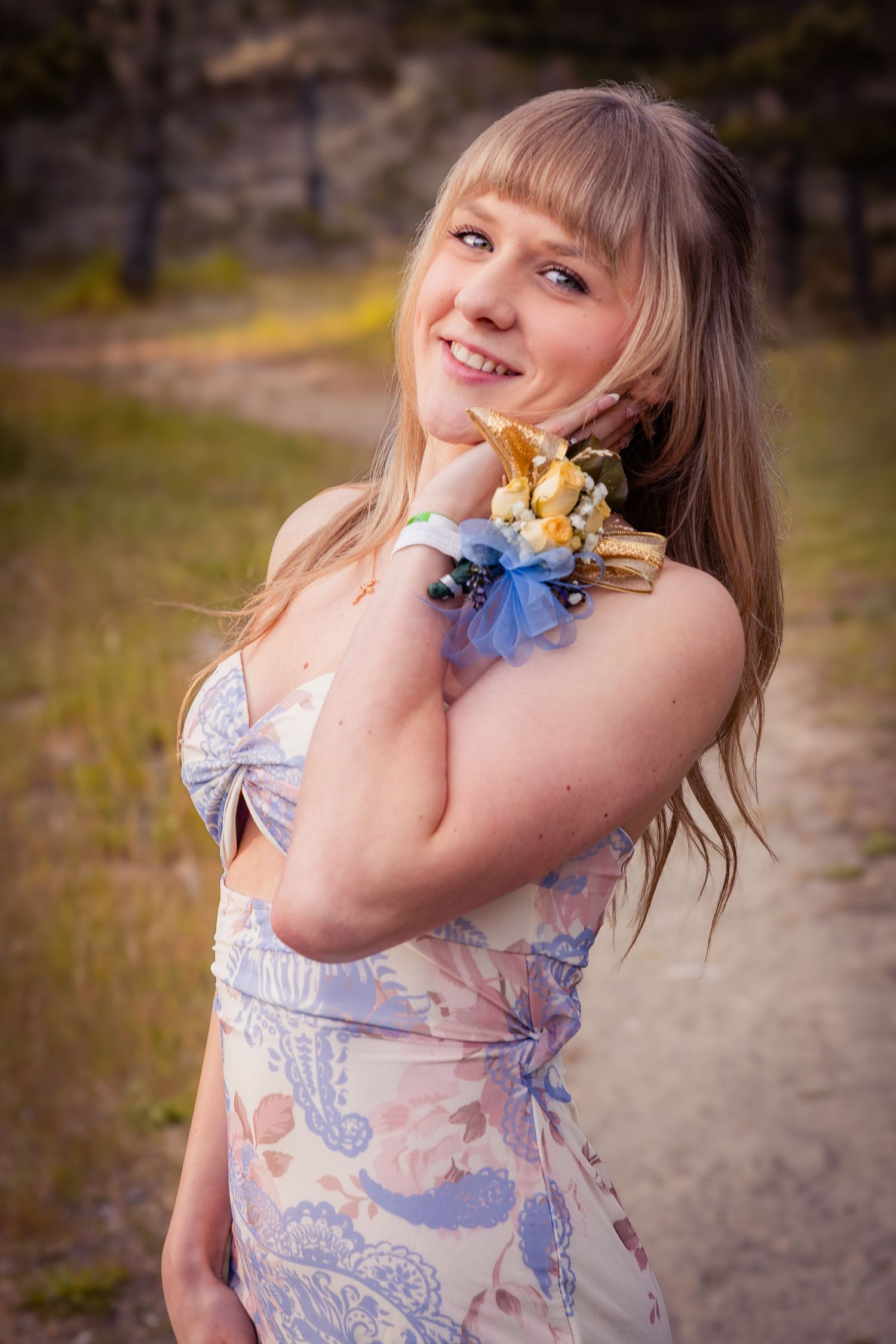 Smiling blonde teen in floral dress displays wrist corsage with blue ribbon outdoors at golden hour.