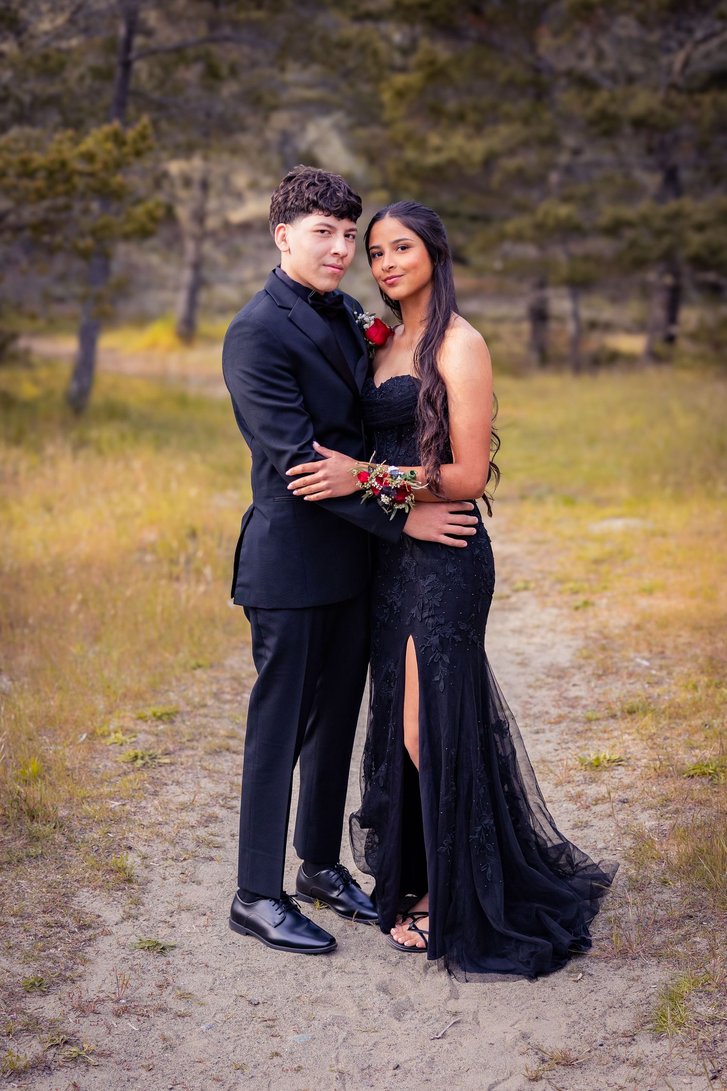Couple in matching black formal wear pose outdoors for prom photos, woman in lace gown and man in tuxedo with red boutonniere.