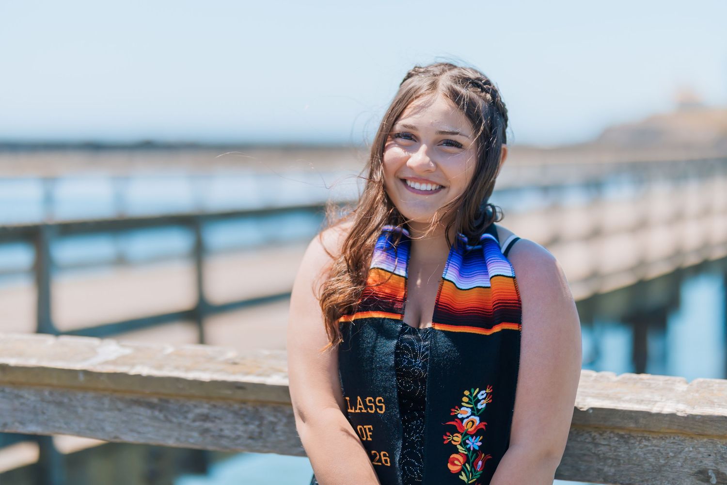 Smiling graduate wearing colorful sash and cap near a sunny waterfront pier on graduation day.
