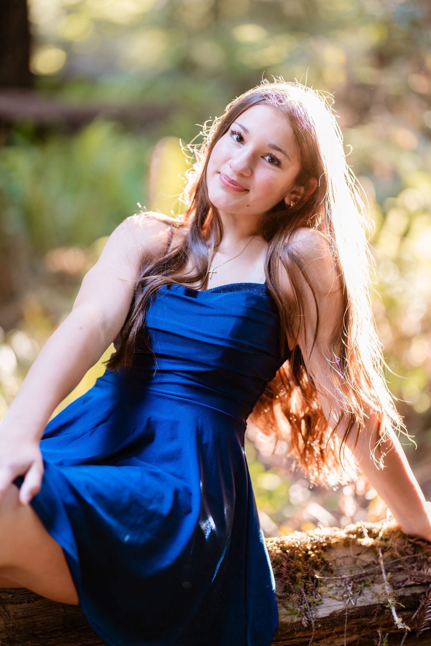 Teen girl in blue dress sitting on log in sunlit forest, long hair flowing, smiling softly.