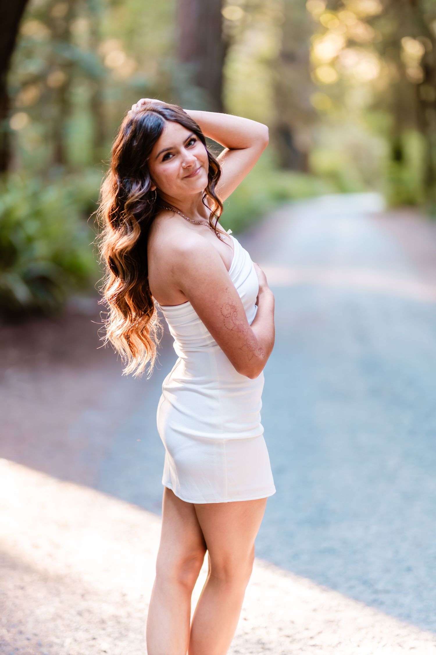 Young woman in white mini dress posing on a sunlit forest path with long wavy hair, smiling over her shoulder.