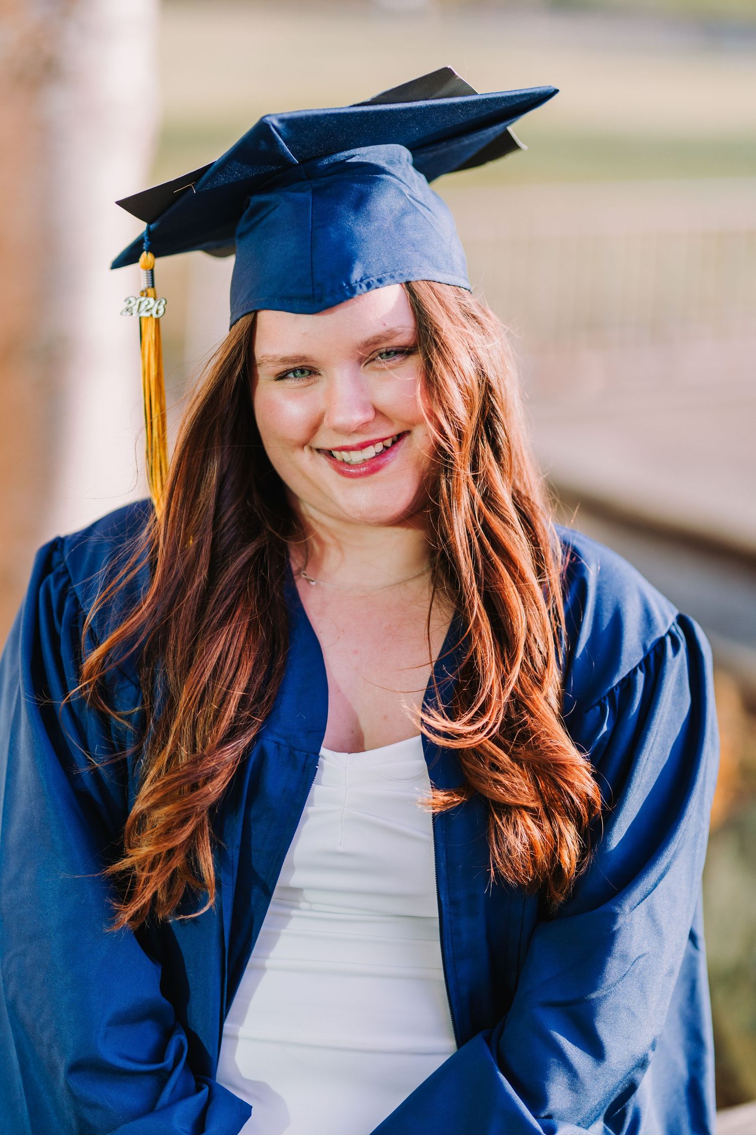 Smiling young woman in blue graduation cap and gown with long red hair posing outdoors for graduation photos.
