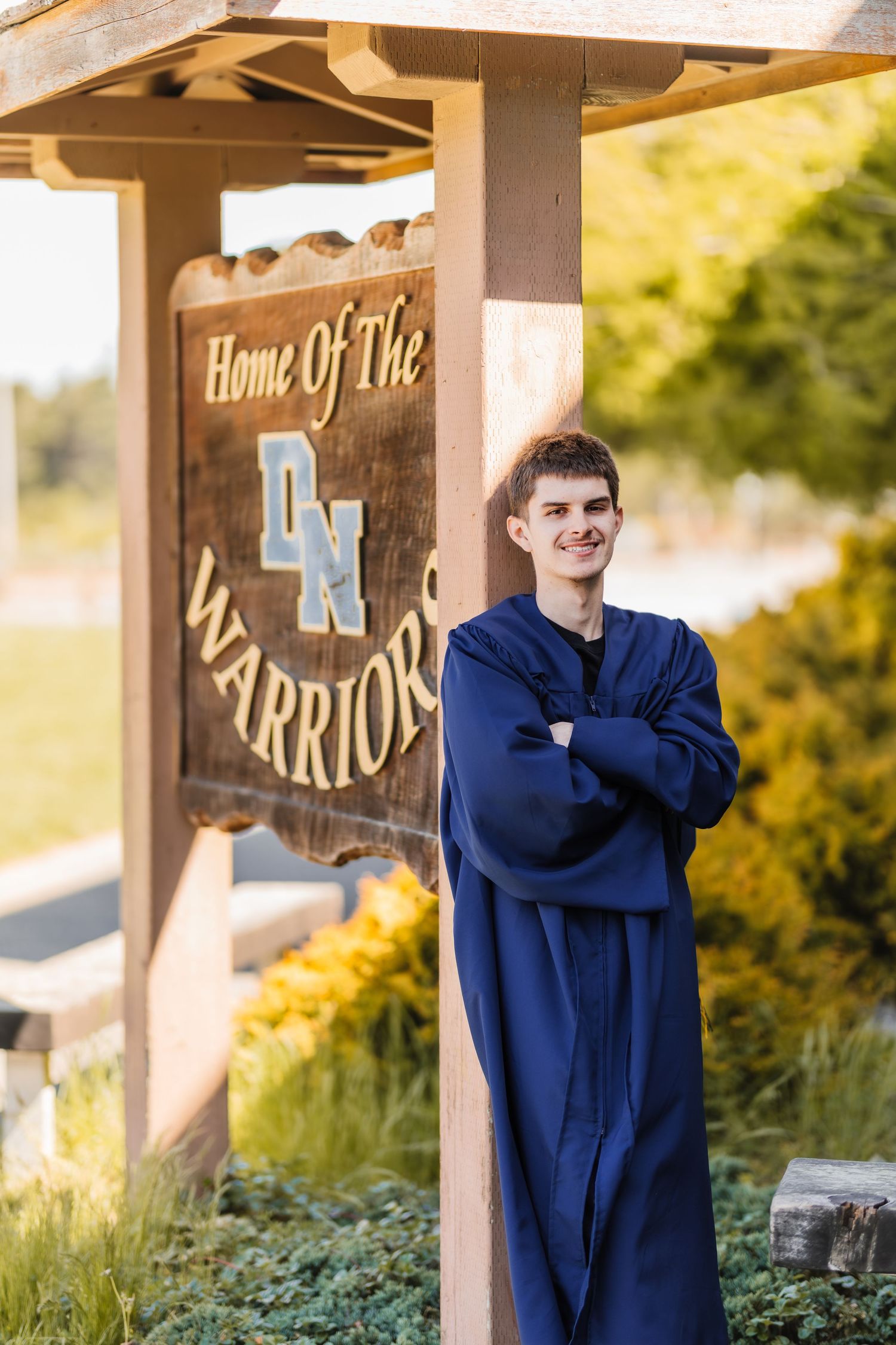 Young man in blue graduation gown stands arms crossed by a Home of the Warriors school sign outdoors.