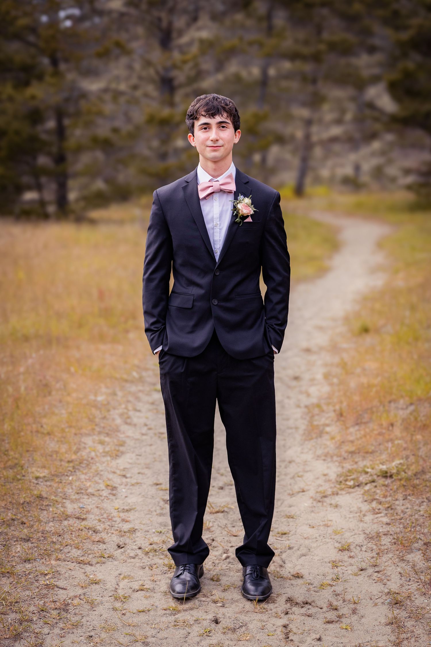 Young man in black suit with pink bow tie and boutonniere stands on dirt path surrounded by autumn field and trees.