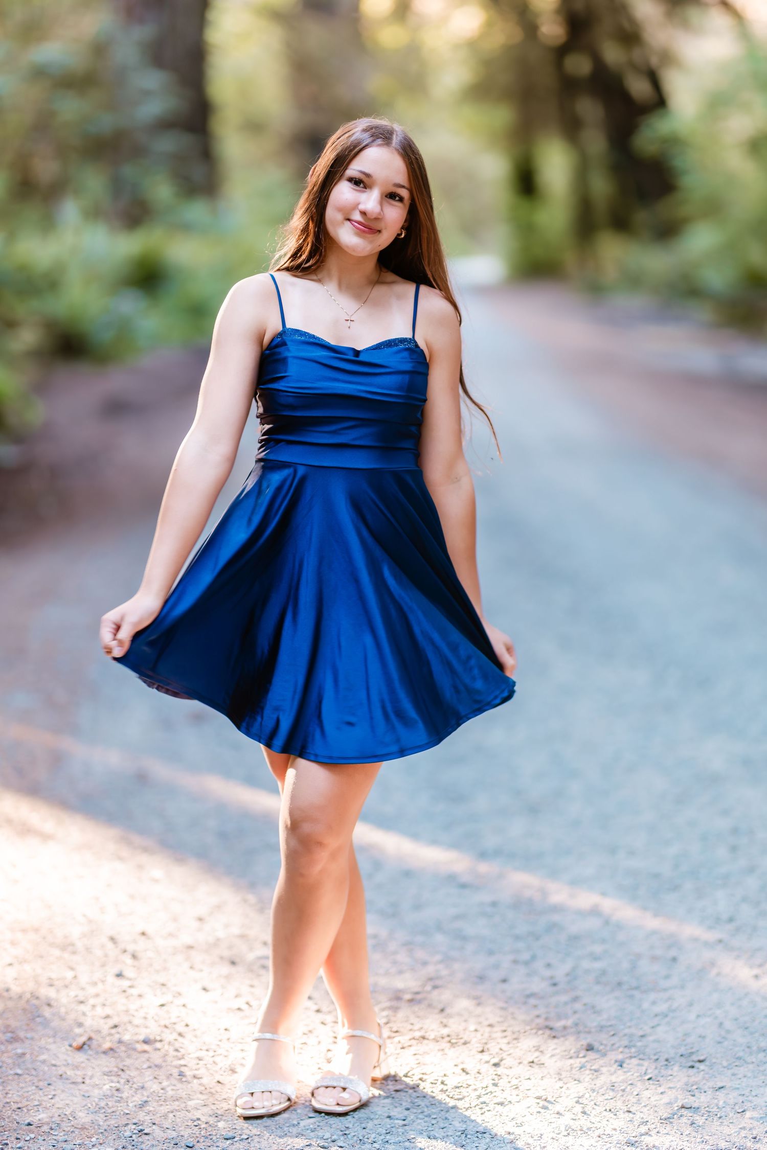Teen girl in royal blue satin dress twirls on a shaded redwood forest road in golden afternoon light.