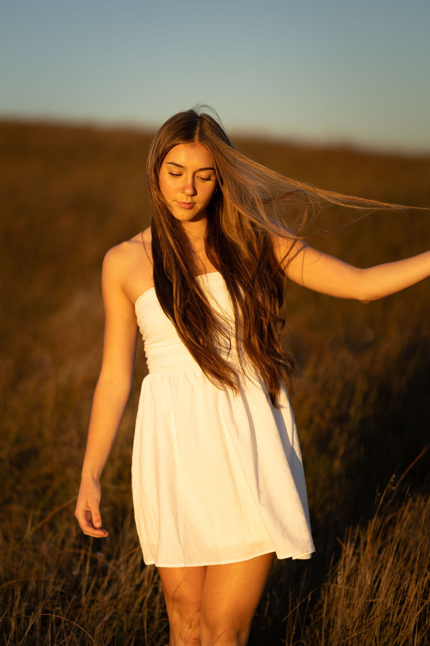 Young woman in white sundress stands in golden field at sunset with long hair blowing in breeze.