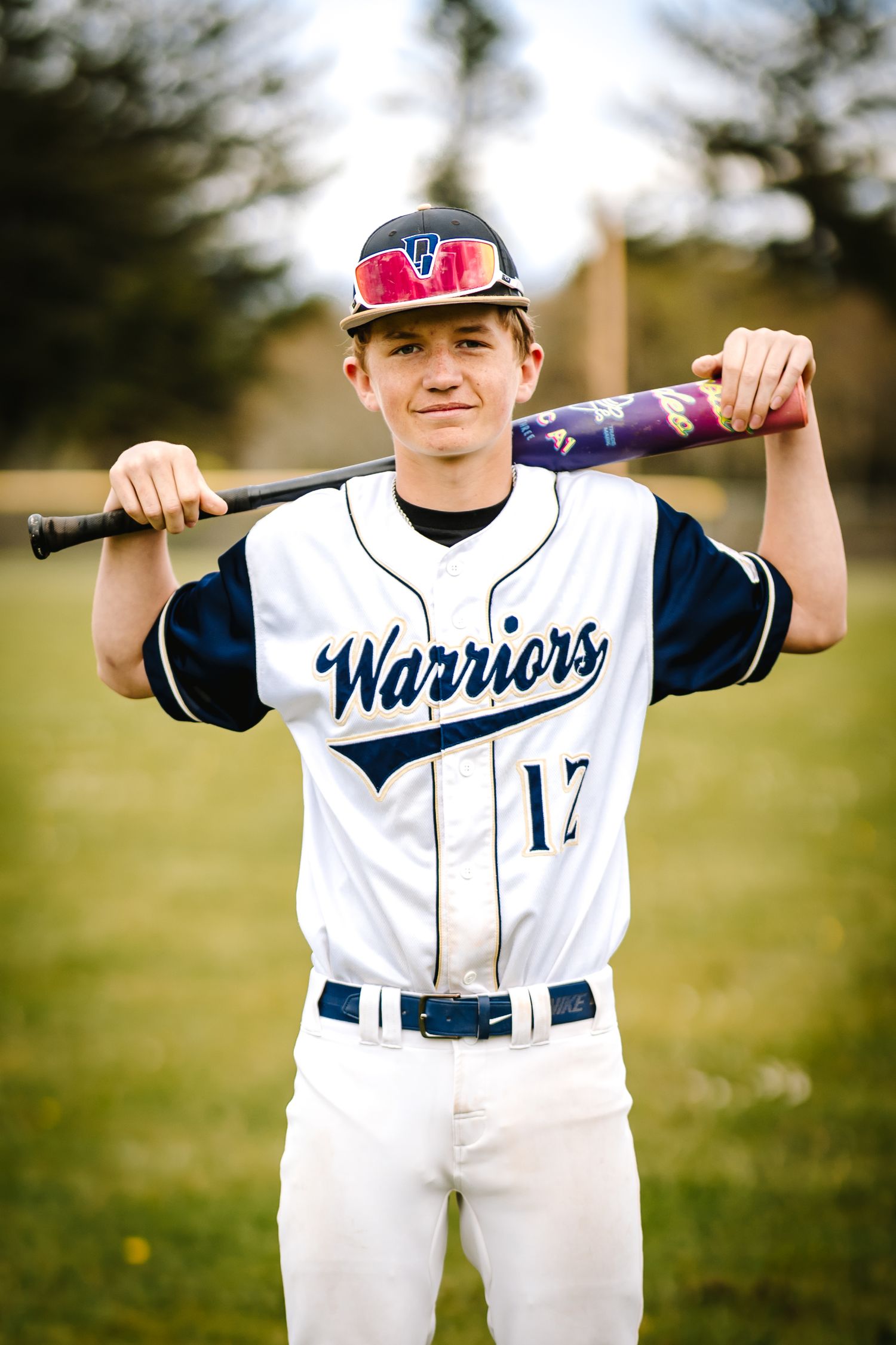 Young baseball player in Warriors number 17 white uniform holding bat across shoulders on a green field.