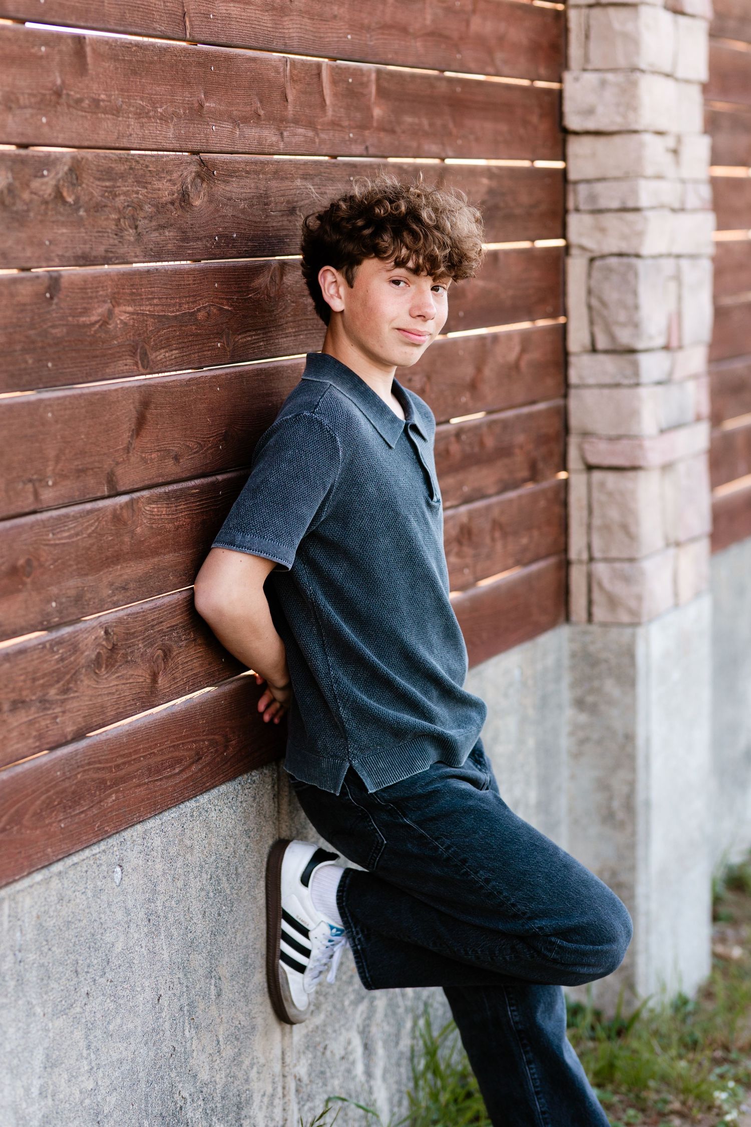 A teenage boy with curly hair leans against a wooden wall, wearing a gray polo shirt, jeans, and Adidas sneakers.