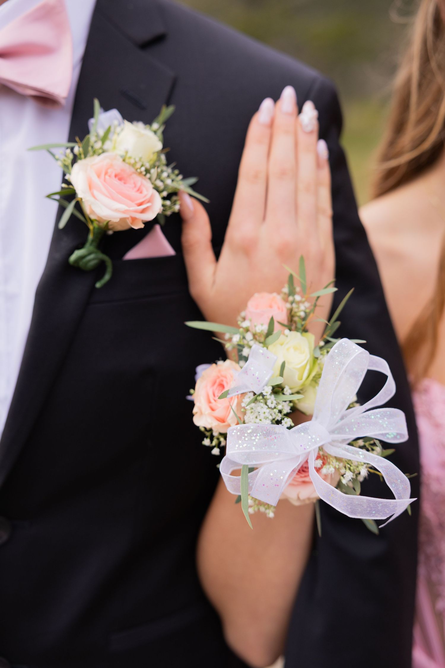 Close-up of a wrist corsage with pink roses and white ribbon bow, paired with a matching boutonniere on a black tuxedo.