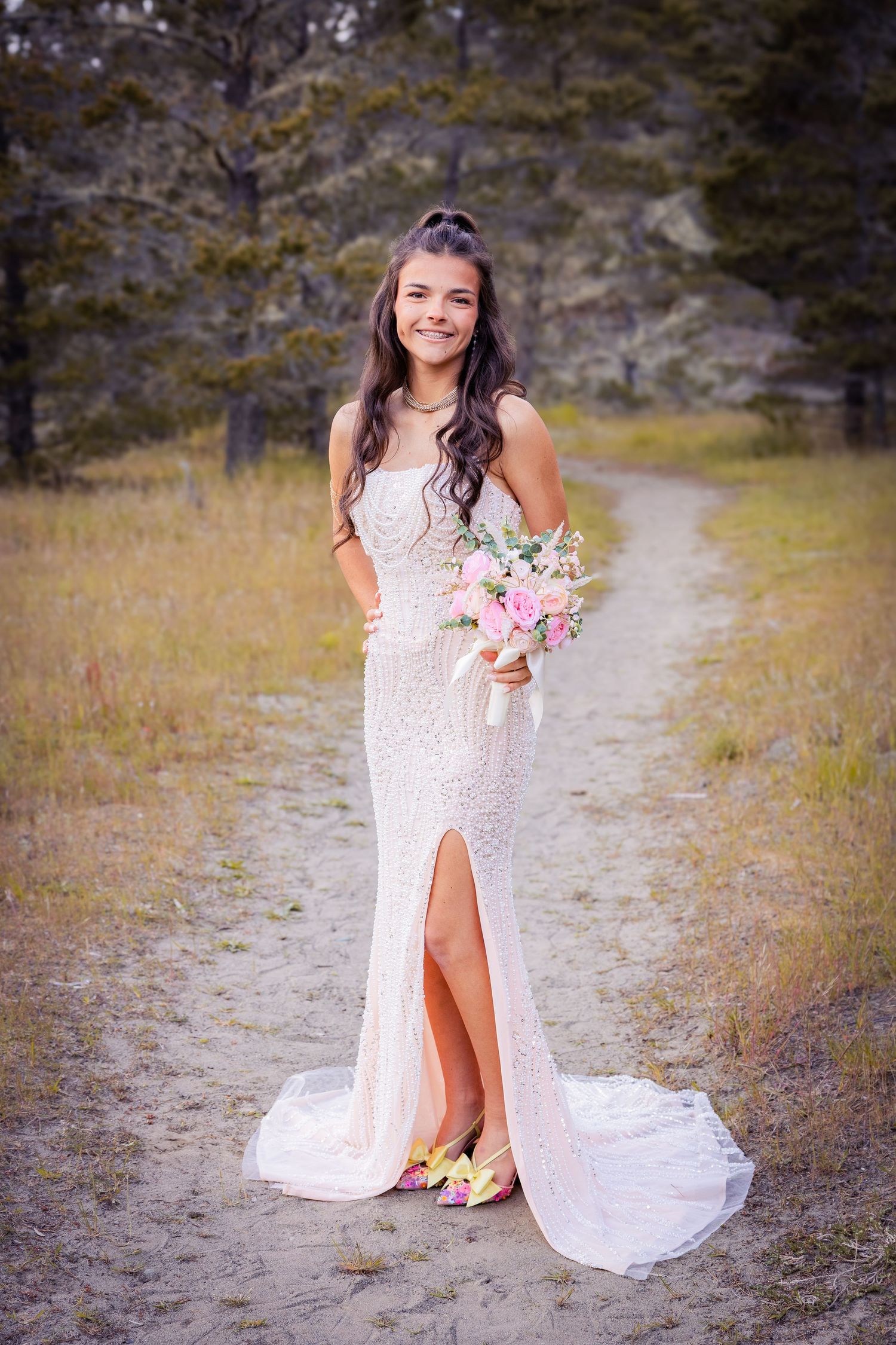 Young bride in white lace gown holding pink bouquet, standing on sandy path surrounded by golden meadow and pine trees.