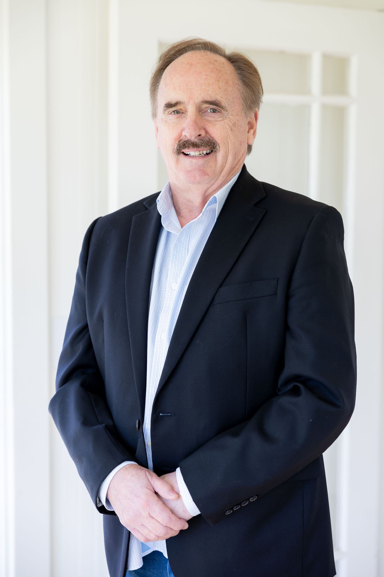 Professional headshot of a smiling middle-aged man with a mustache wearing a dark blazer and light blue shirt.