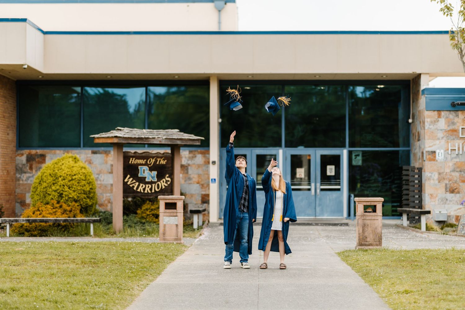 Two graduates in blue gowns celebrate outside their Warriors school building on graduation day.