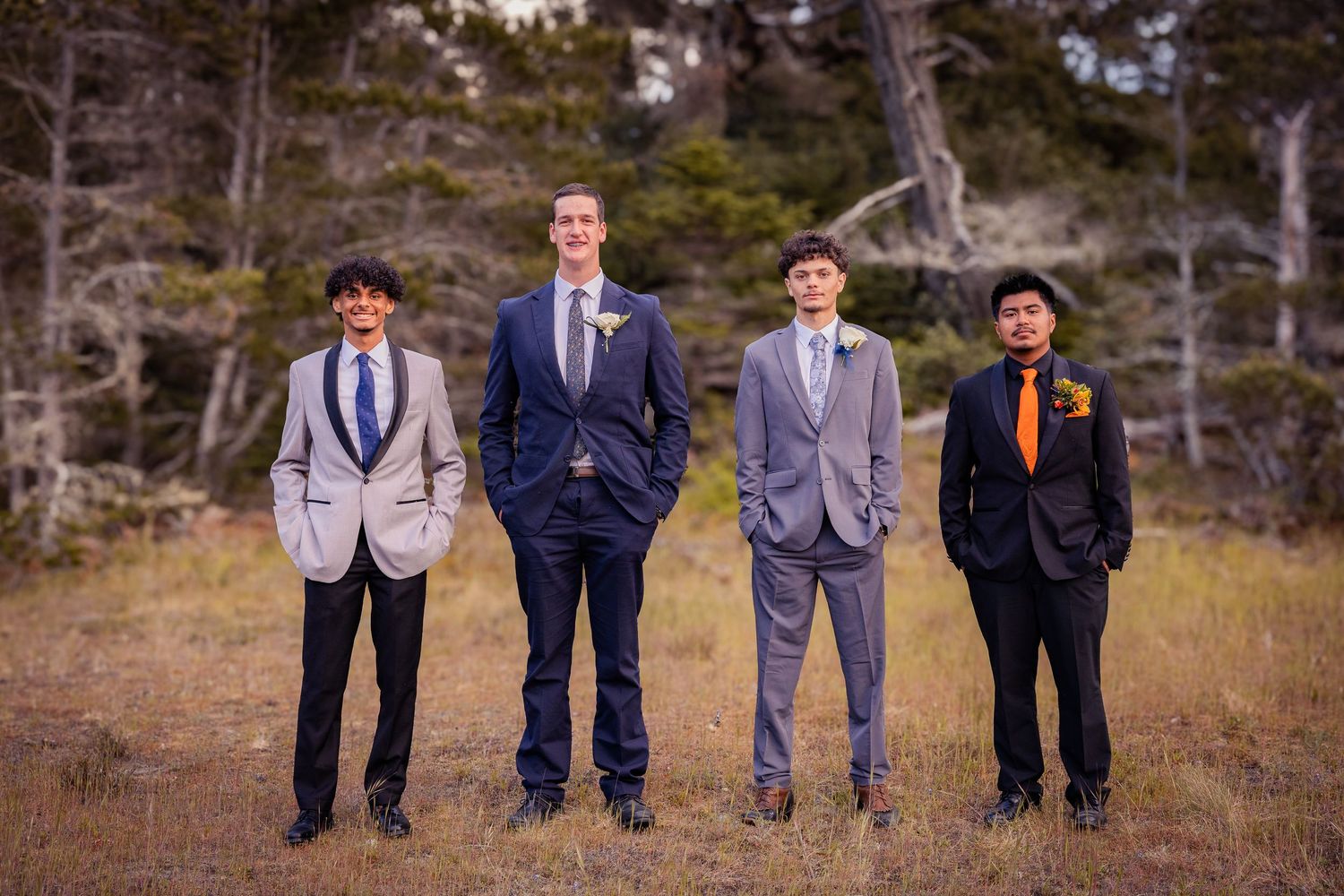 Four groomsmen in suits standing in a field, with trees in the background at an outdoor wedding.