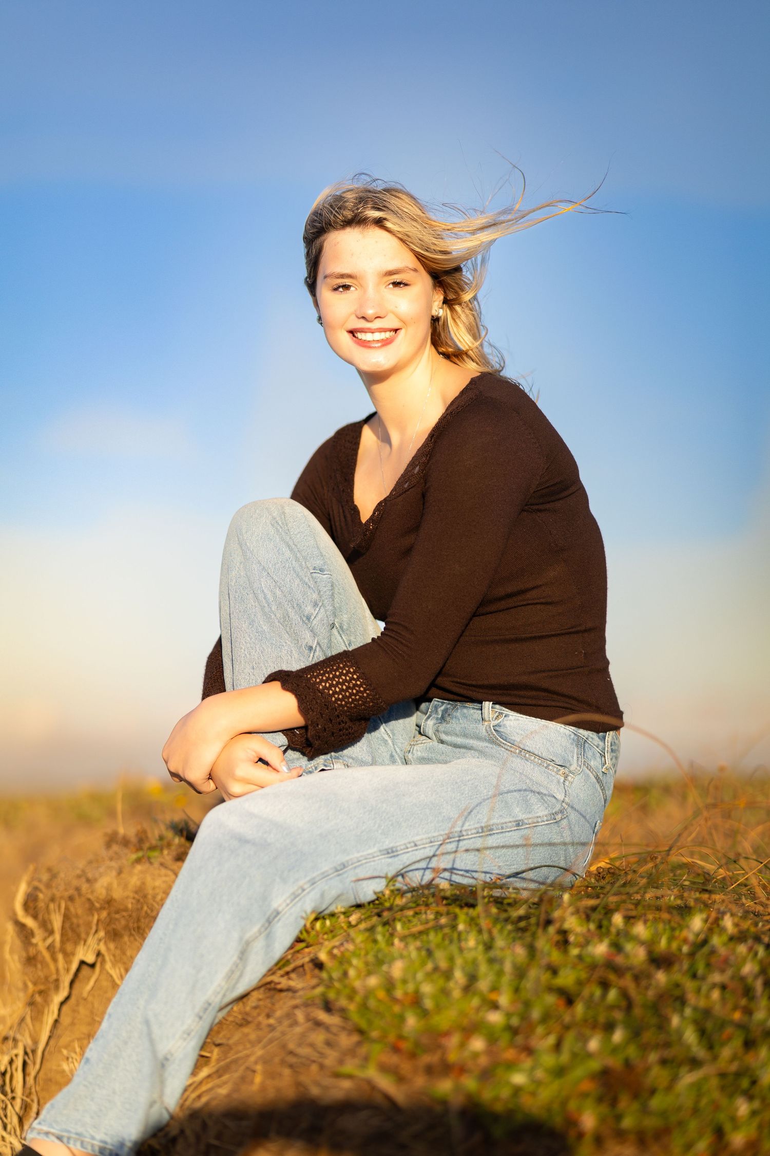 Smiling blonde teen girl sitting outdoors in black long-sleeve top and light jeans under blue sky.