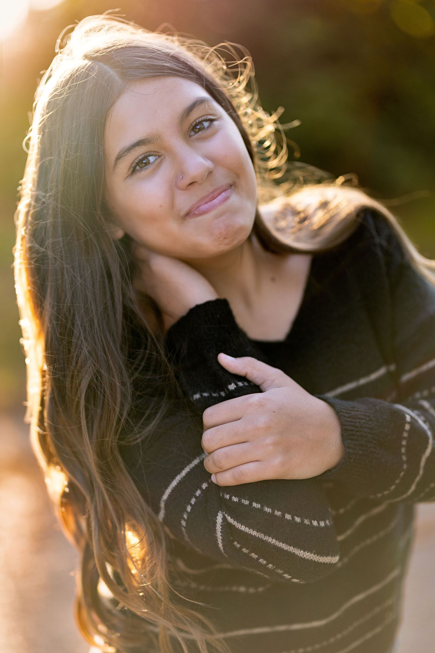 Teenage girl with long dark hair smiling warmly in golden backlit sunlight wearing a black striped sweater.