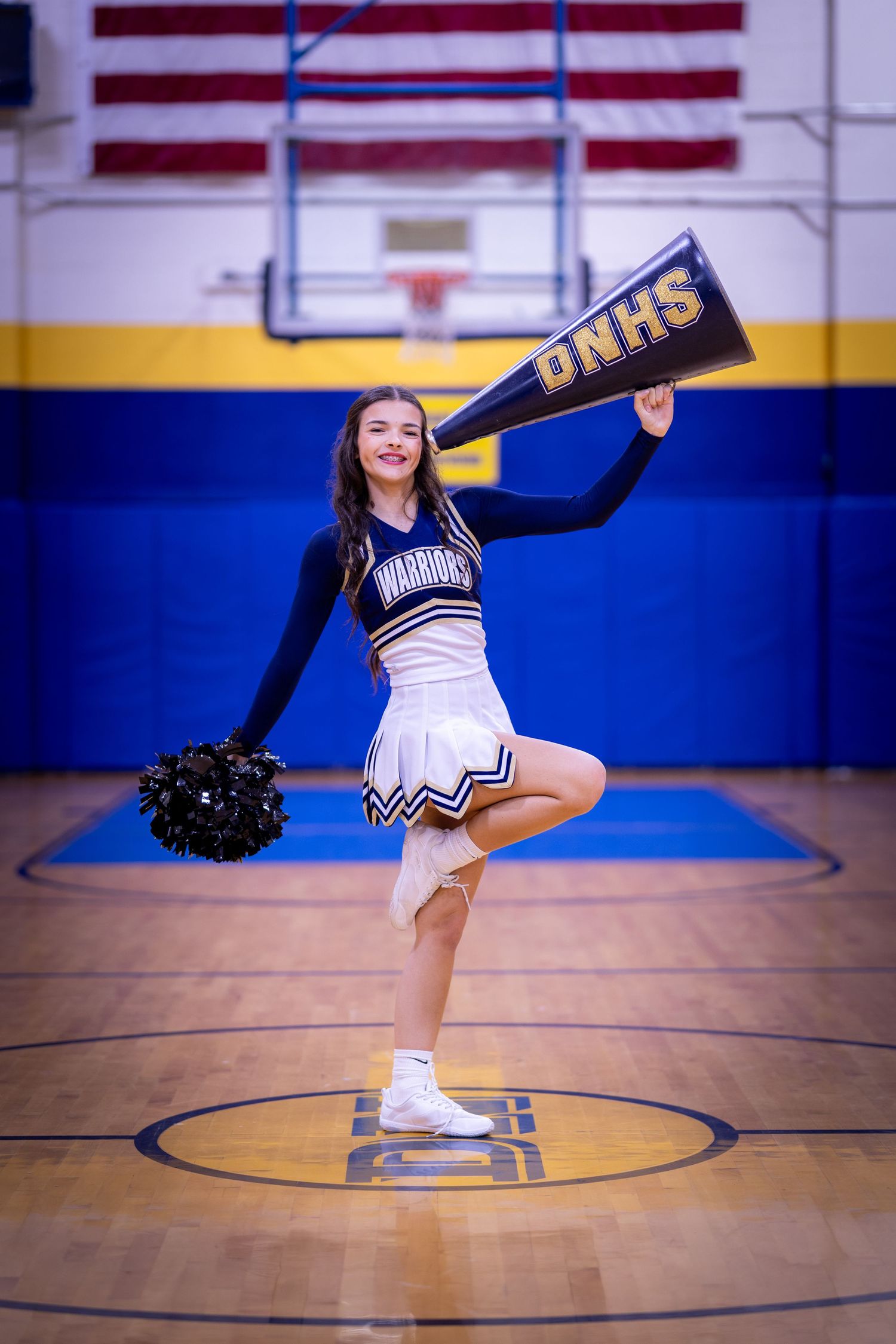 A cheerleader in a Warriors uniform holds a DNHS pennant while posing on one leg in a school gymnasium.