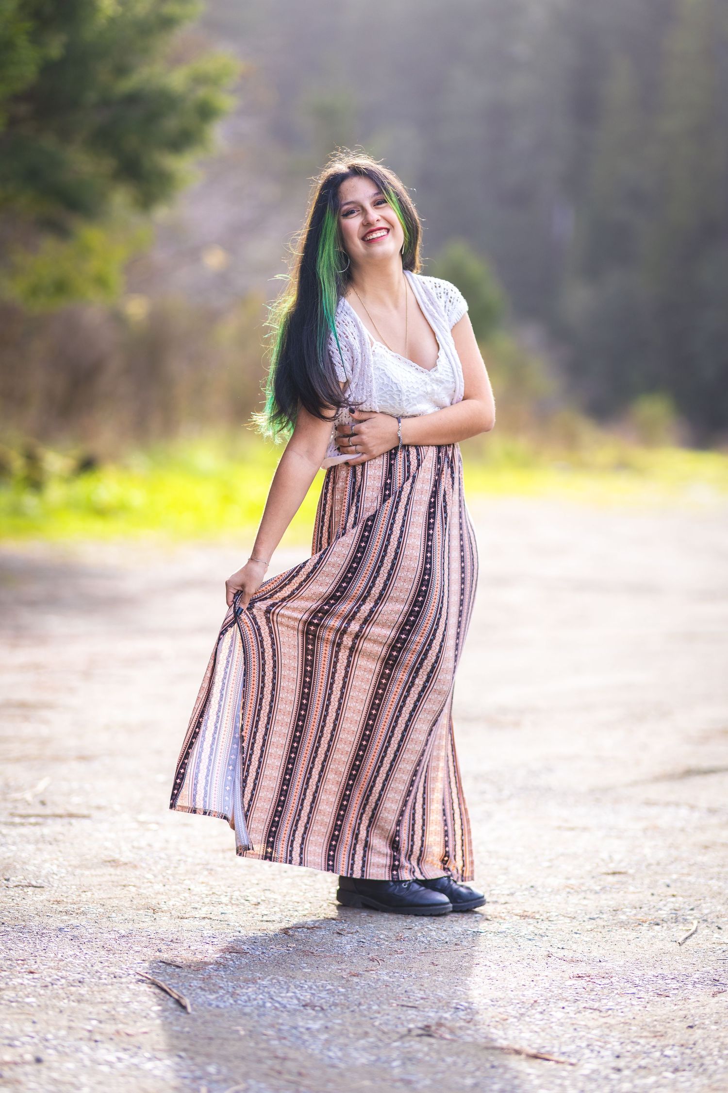 Smiling young woman with green-highlighted hair twirls a striped maxi skirt on a sunlit path.