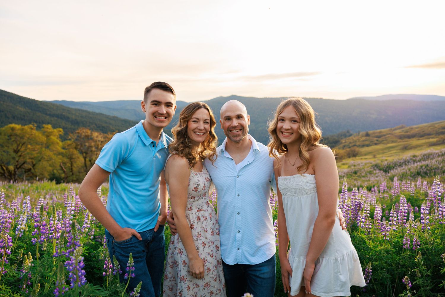 Happy family of four smiling together in a field of purple lupine flowers at sunset.