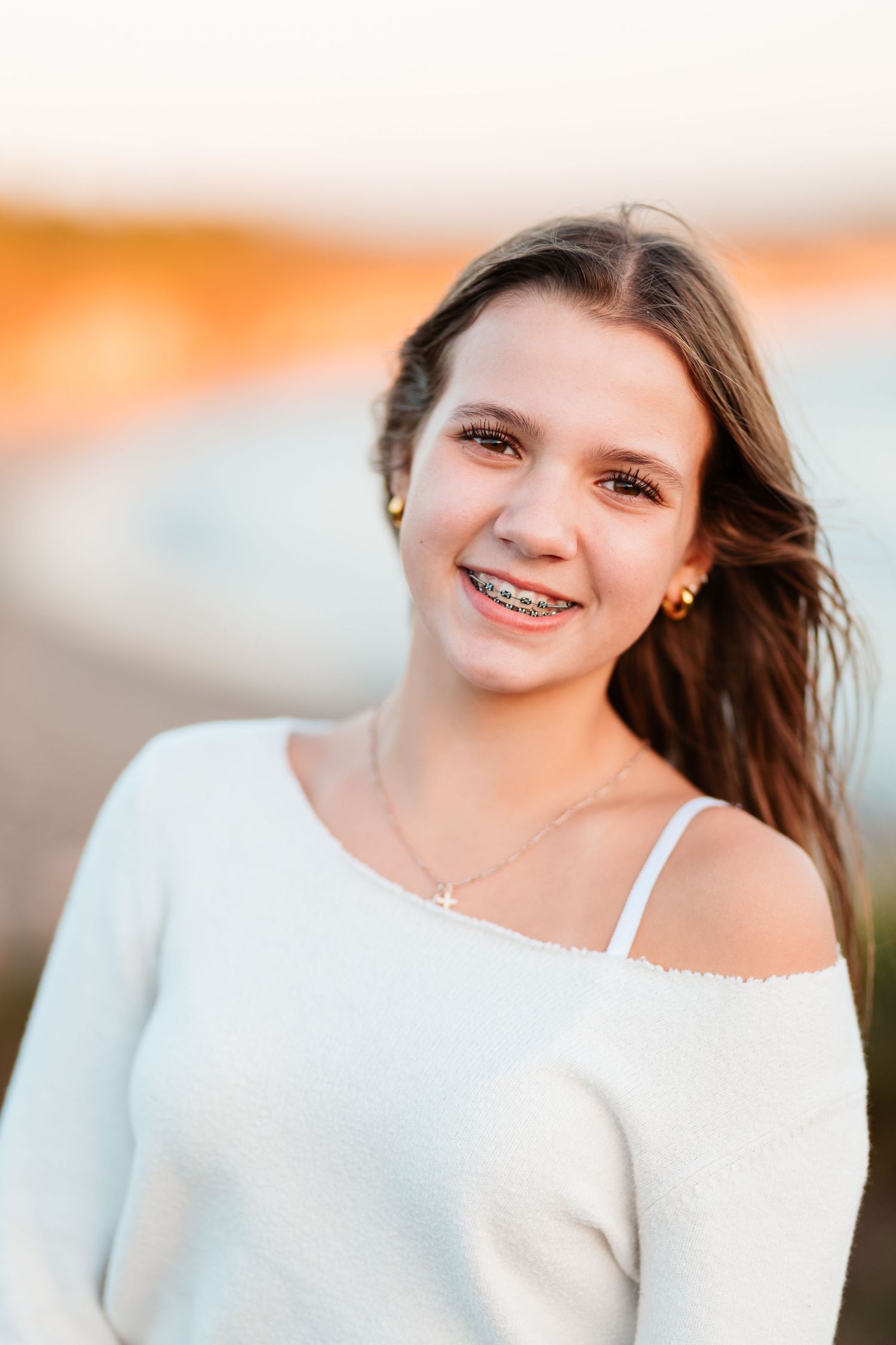 Smiling teenage girl with braces wearing a white off-shoulder sweater in warm sunset light outdoors.