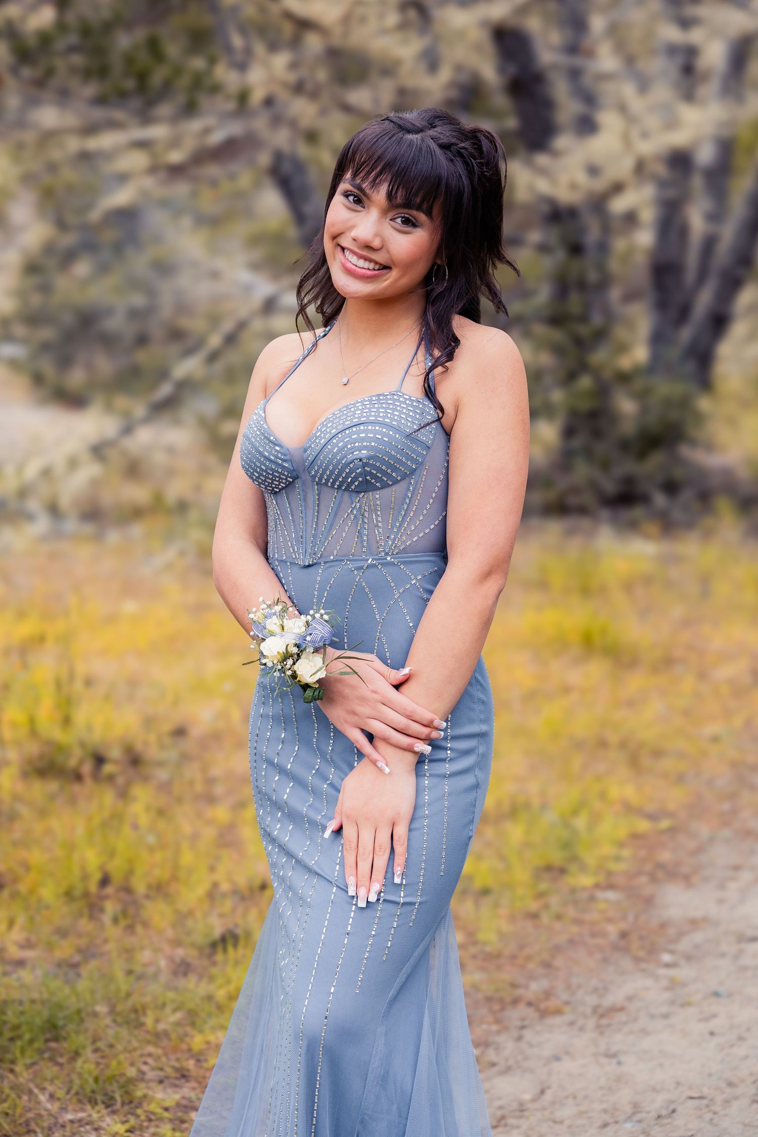 Young woman posing in a stunning blue mermaid prom dress with rhinestone details and a floral wrist corsage.