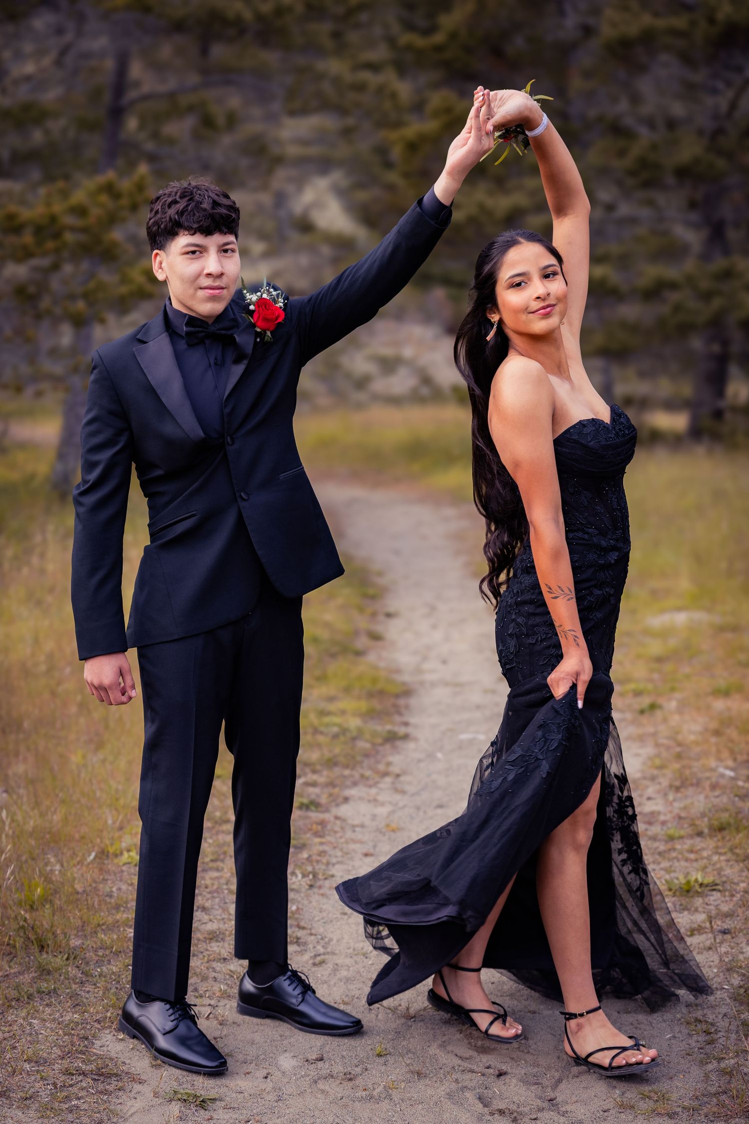 A young couple in matching black formal wear dance outdoors, he in a suit with red boutonniere, she in a strapless gown.