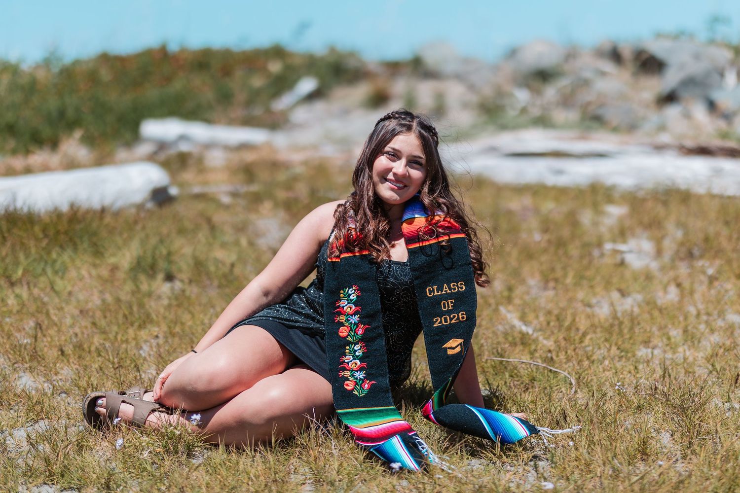 Young woman sitting outdoors in graduation attire holding a Class of 2020 mortarboard in a grassy field.