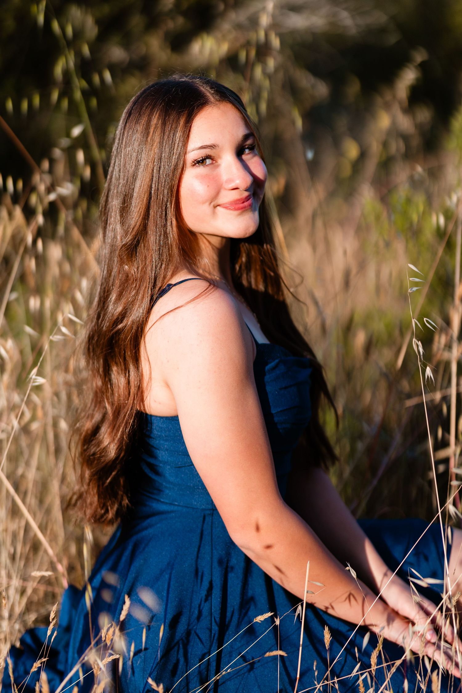 Young woman in blue dress sitting in golden field grass, long brown hair, smiling softly in warm sunlight.