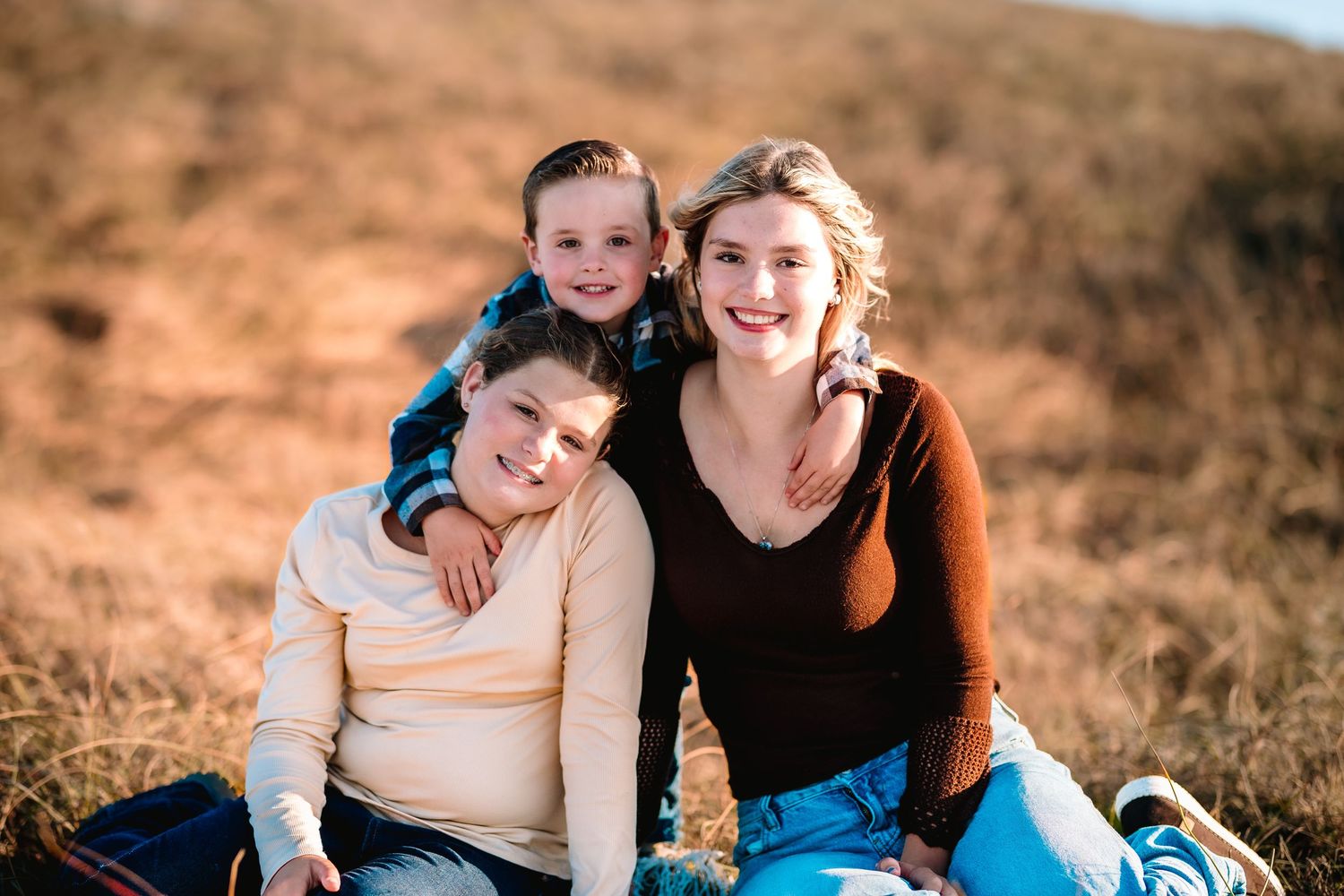 Three smiling people pose together in a golden field, with two seated and one child standing behind them.