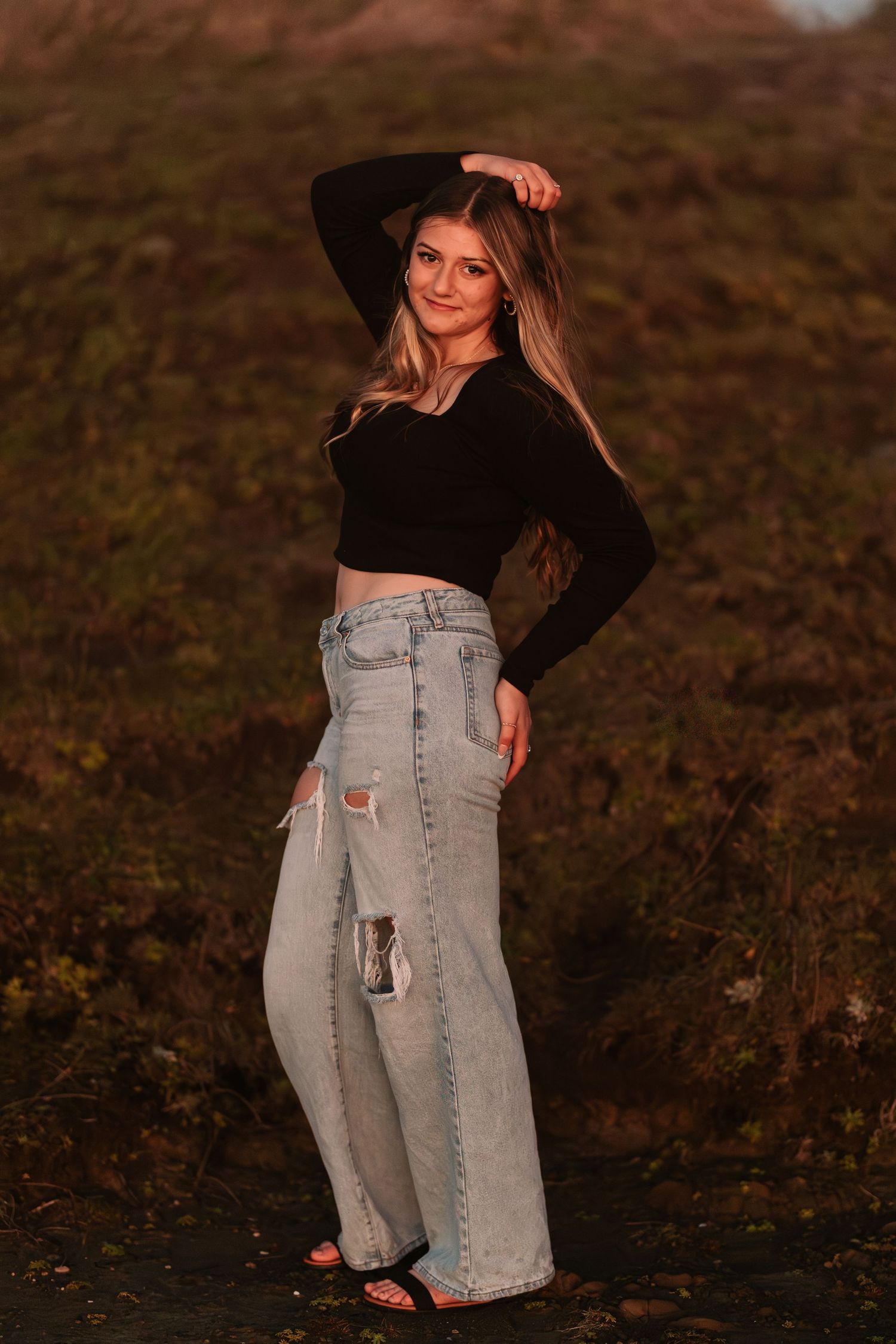 Teen girl in black crop top and distressed wide-leg jeans posing outdoors during golden hour.