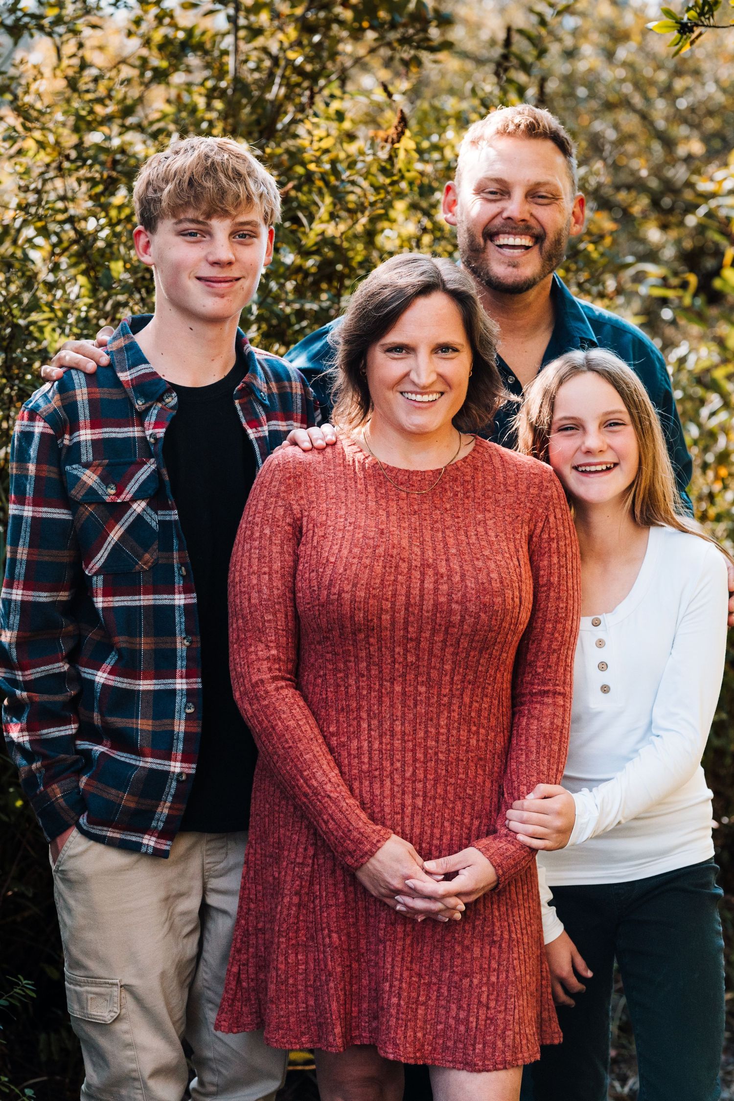 Happy family of four smiling outdoors in fall, wearing casual clothes with green foliage in background.