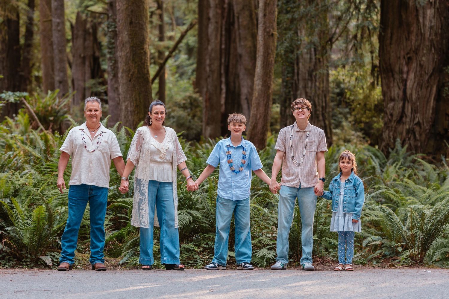 Family of five holding hands on a forest path surrounded by lush ferns and tall trees.