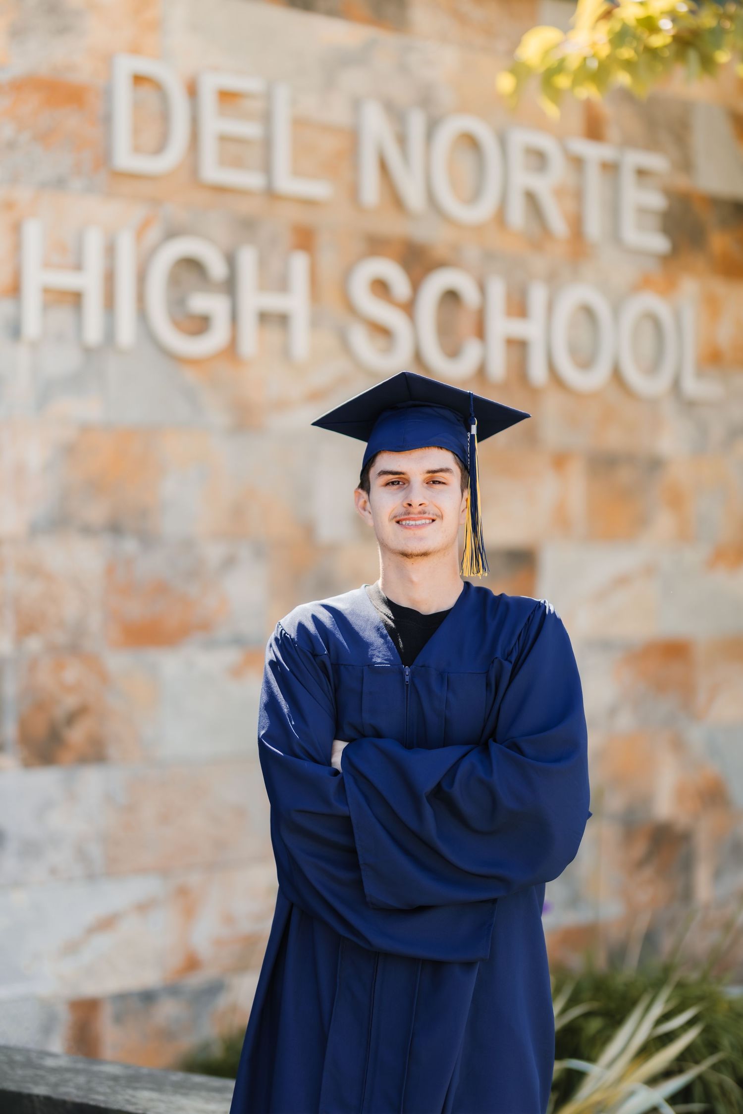 A smiling graduate in blue cap and gown stands arms crossed in front of Del Norte High School stone sign.