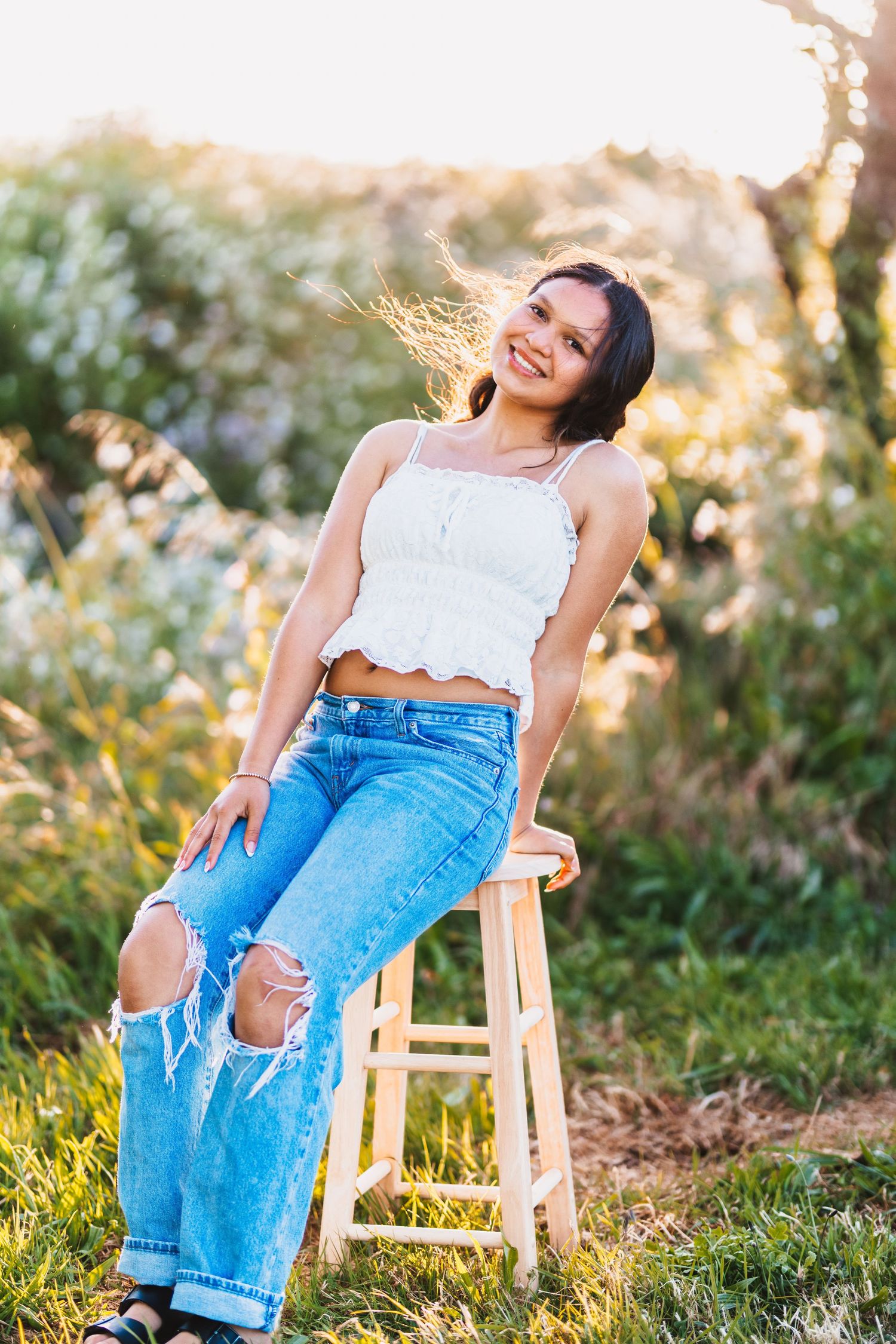 Young woman in white crop top and ripped jeans sitting on stool outdoors in golden sunlight.