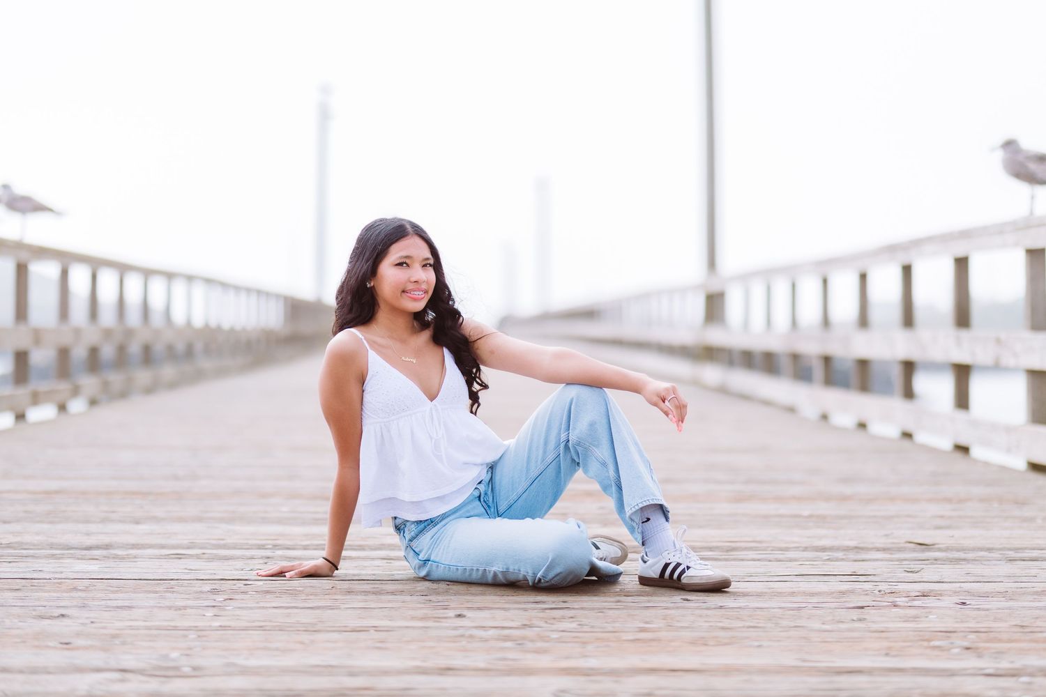 Young woman in white top and jeans sitting on a wooden pier, smiling confidently on a misty day.