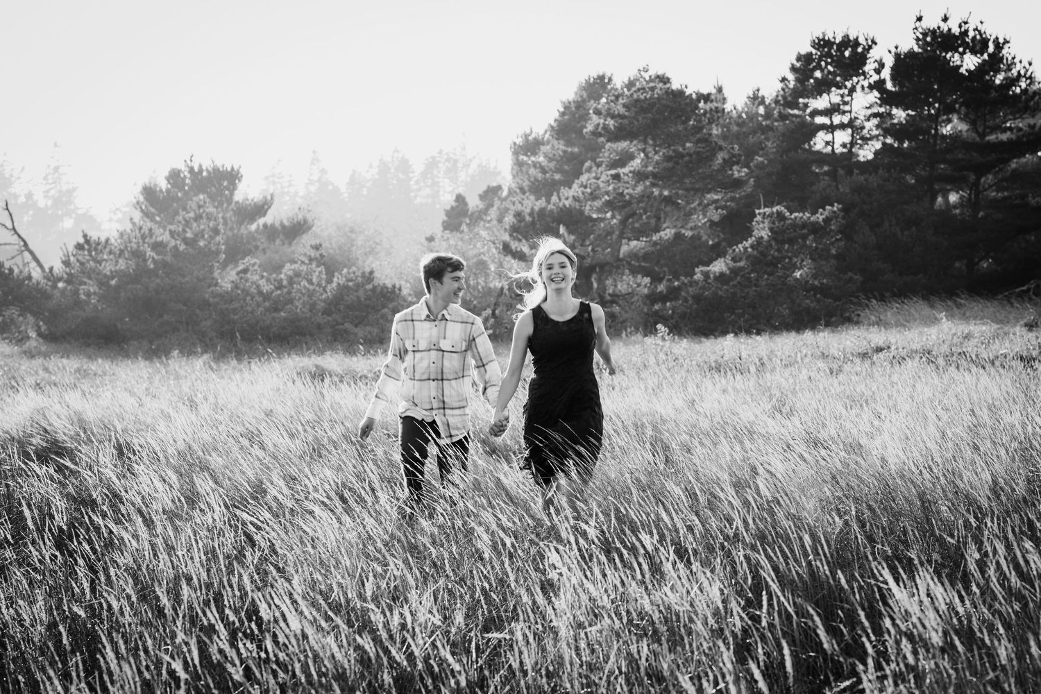 Black and white photo of a couple holding hands walking through tall grass field with trees in background.