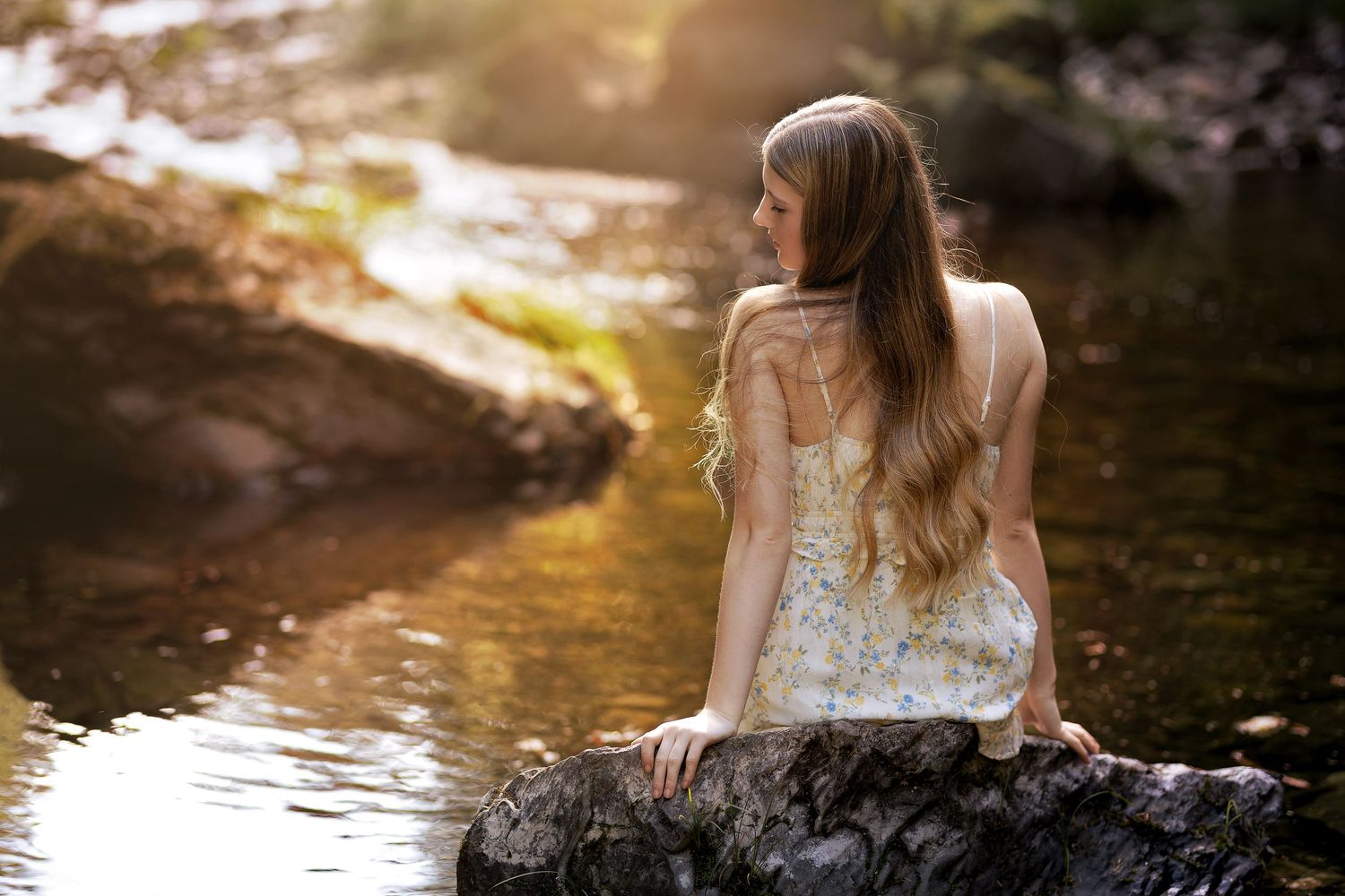Young woman with long wavy hair in a floral dress sitting on a rock by a sunlit stream.