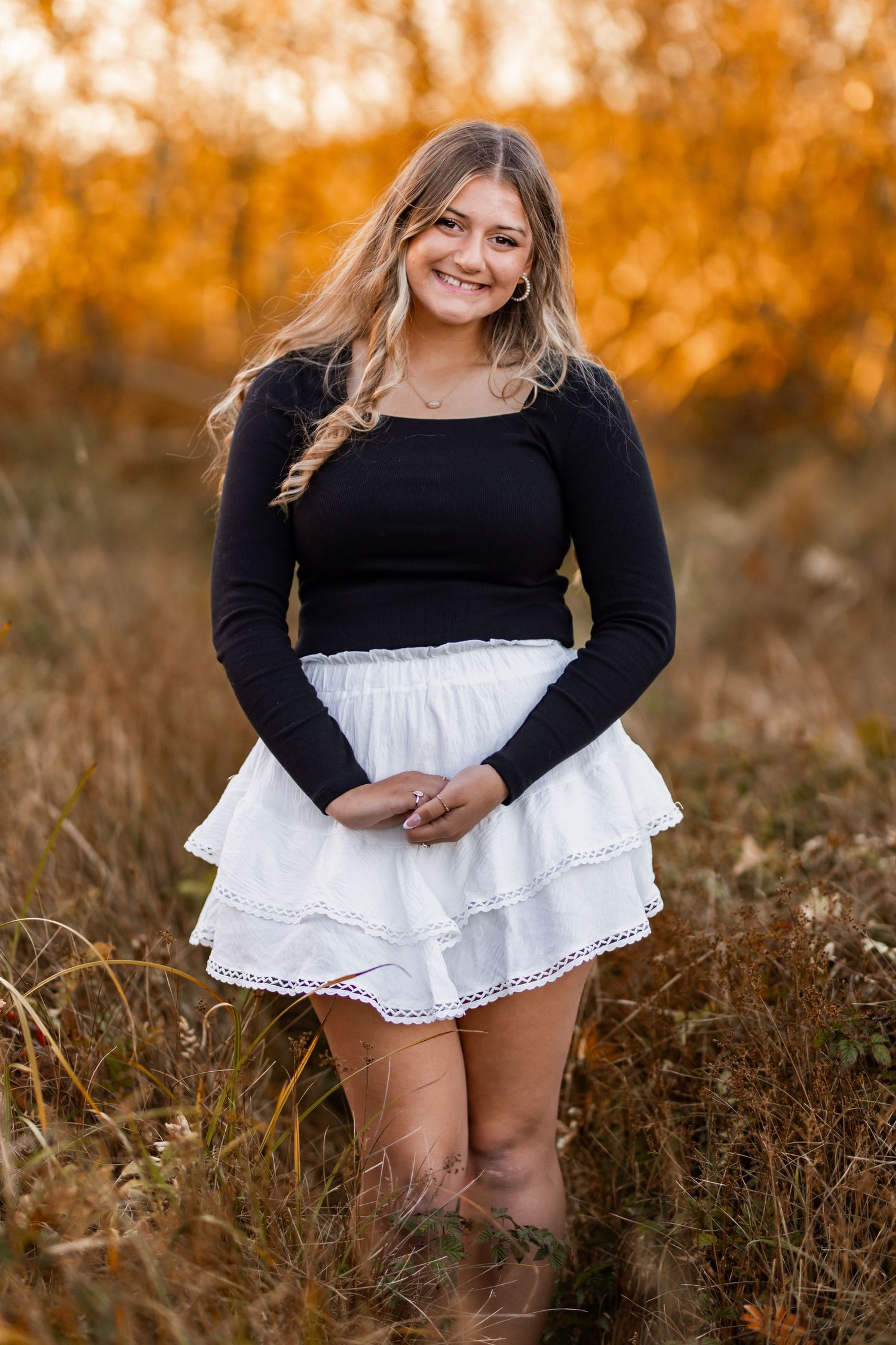 Smiling young woman in black long-sleeve top and white tiered skirt standing in an autumn field.