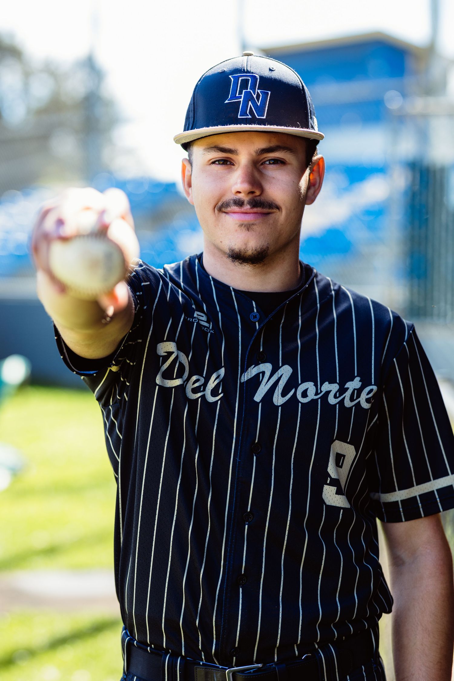 Del Norte baseball player in pinstripe uniform extending baseball toward camera on sunny field.