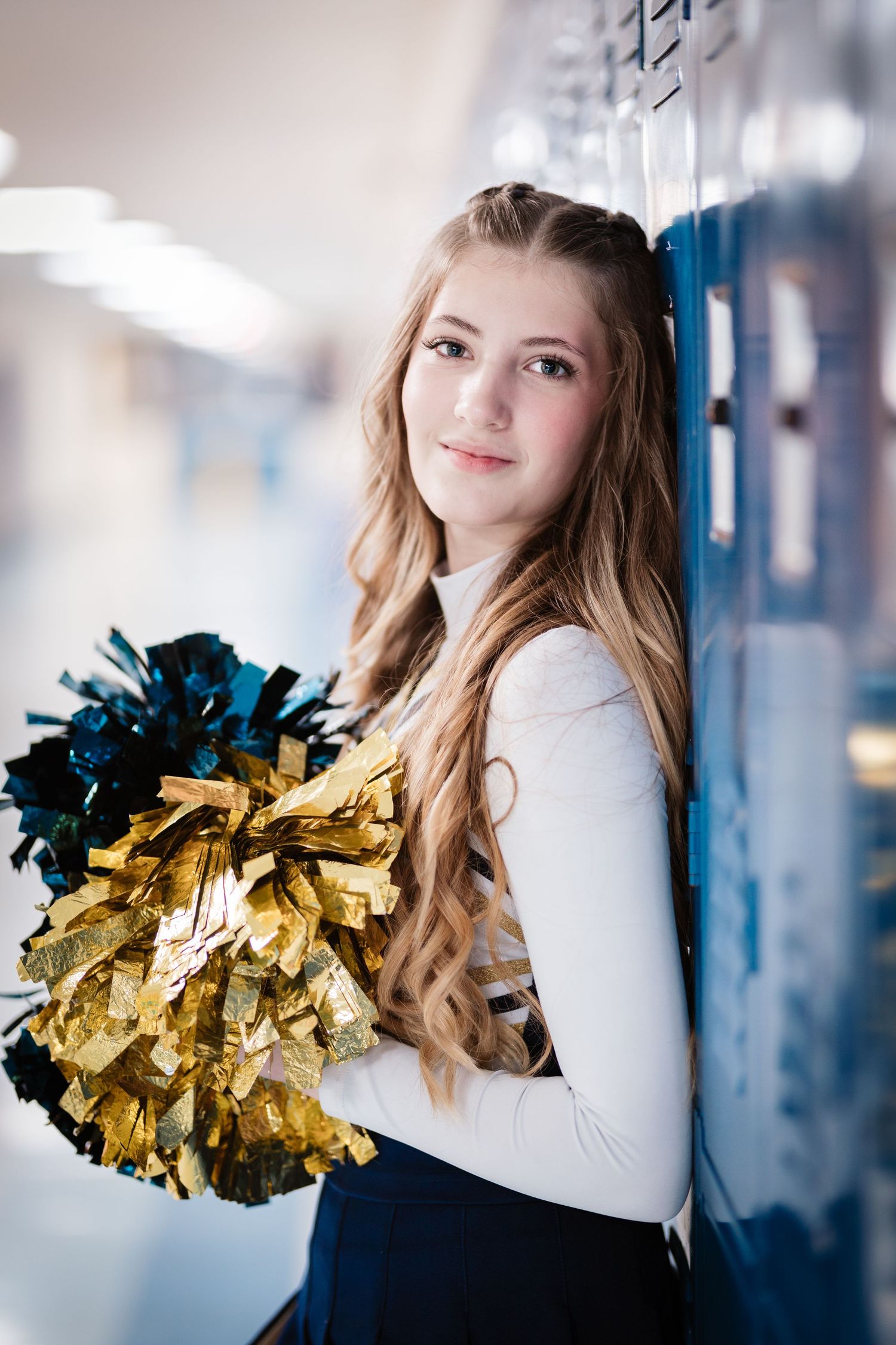 Blonde cheerleader leans against blue lockers holding gold and blue pom-poms.