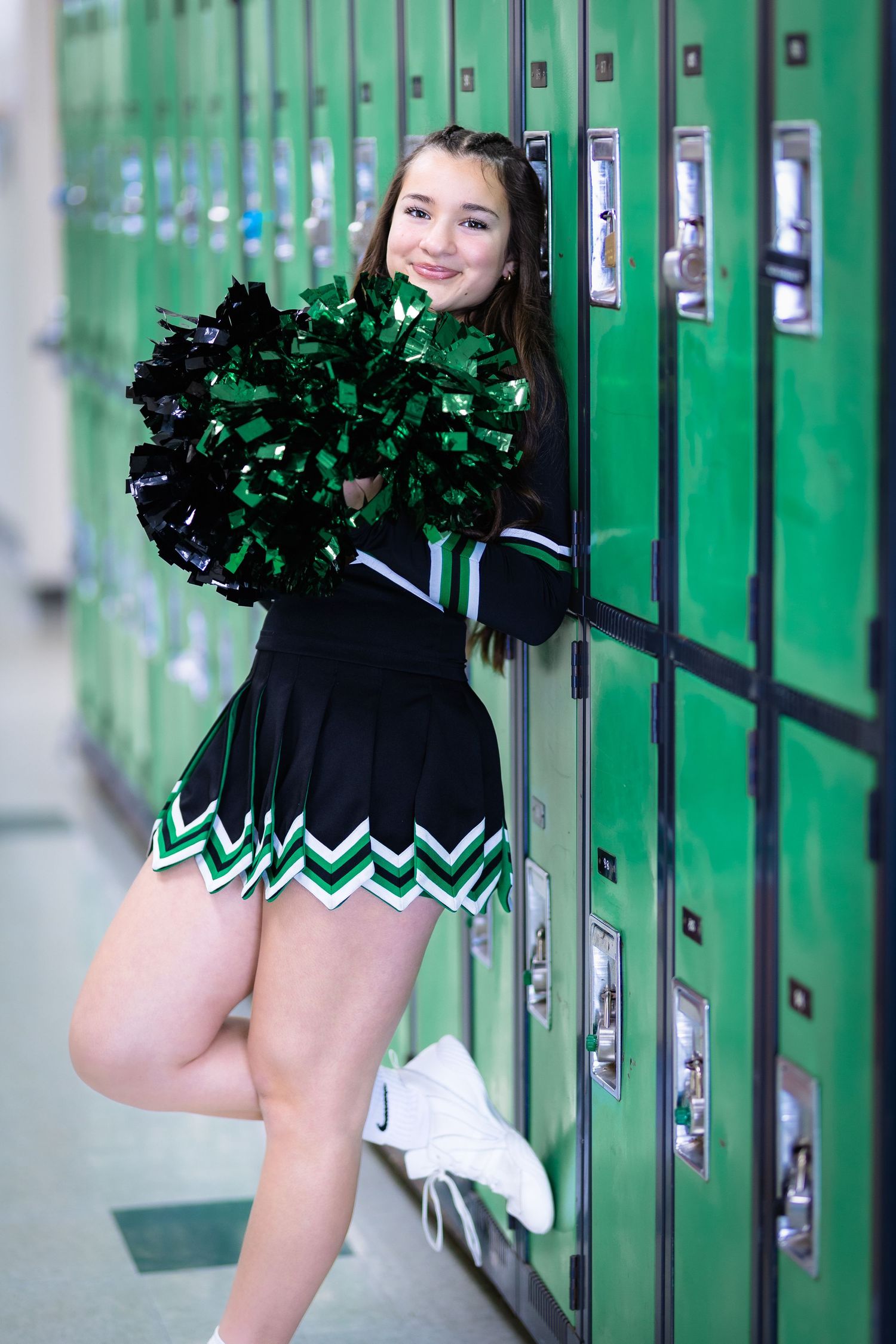 A cheerleader in a black and green uniform leans against green lockers holding pom-poms.