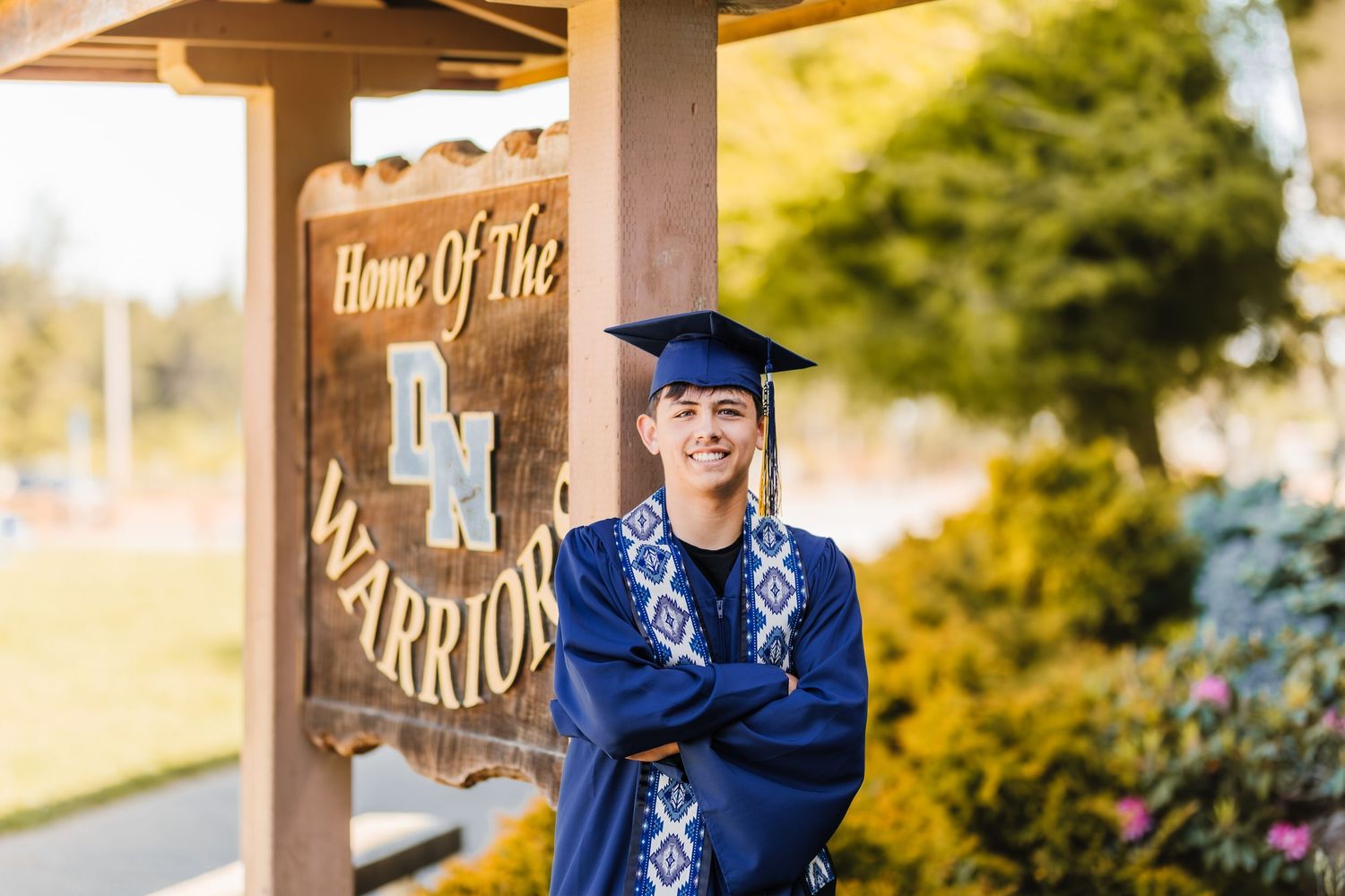 Smiling male graduate in blue cap and gown stands arms crossed by a Warriors school sign outdoors.