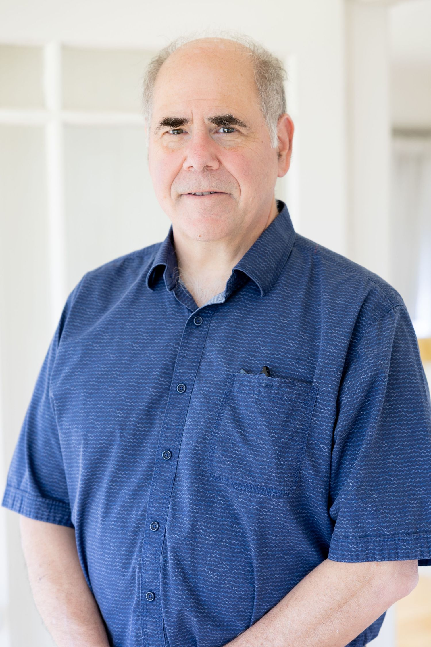 Middle-aged man with gray hair wearing a blue short-sleeve button-up shirt, smiling indoors in bright natural light.