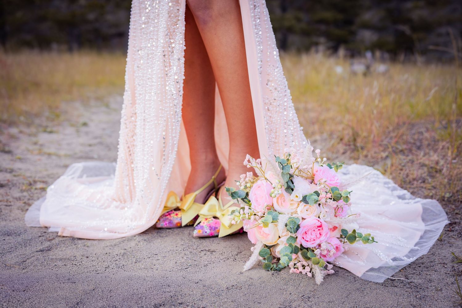 Bride in sequined gown with colorful heels and pink floral bouquet on sandy ground.