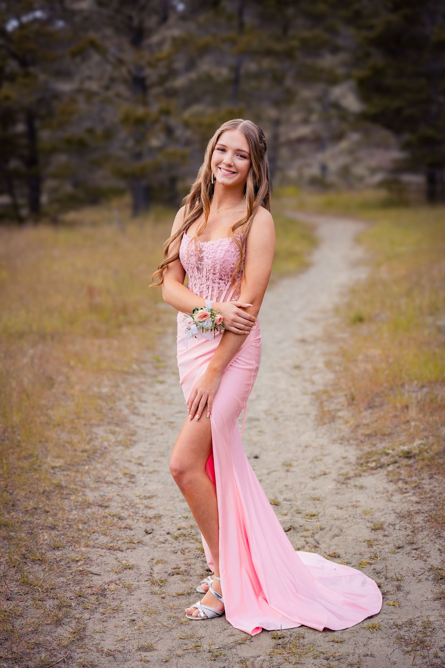 Young woman in pink sequin prom gown with floral corsage poses on dirt path surrounded by trees.