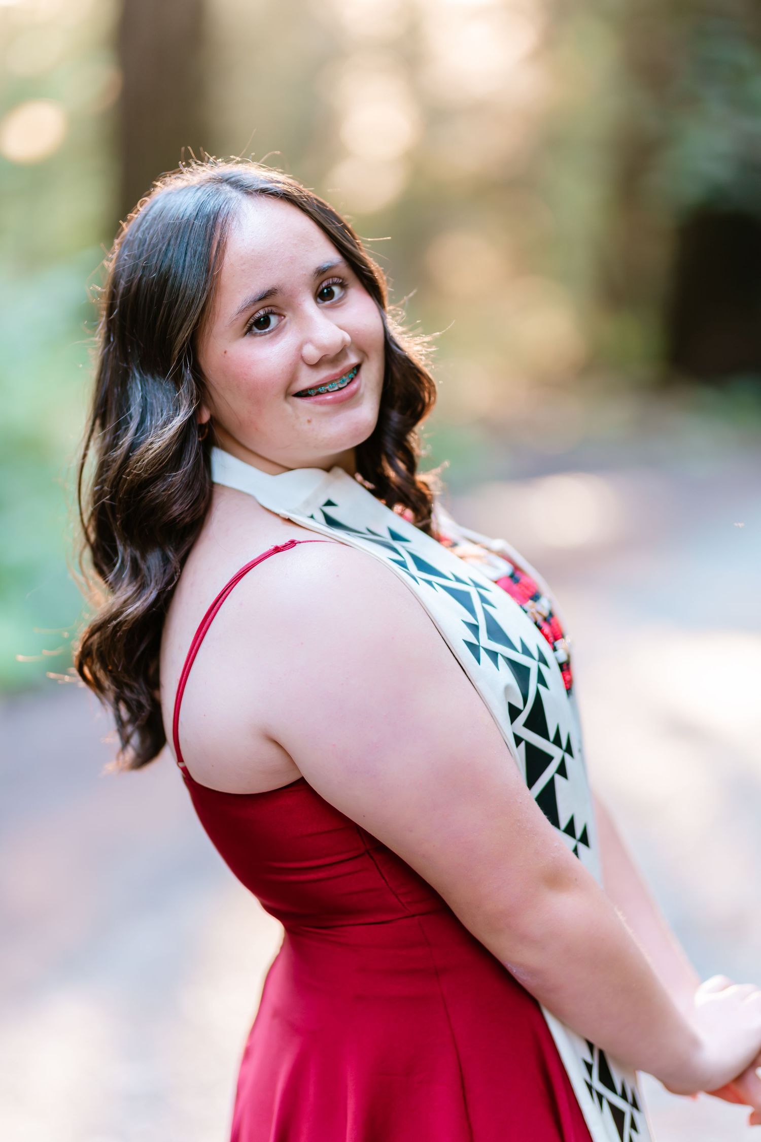 Young woman with braces in red dress and geometric sash smiles over shoulder on sunlit forest path.