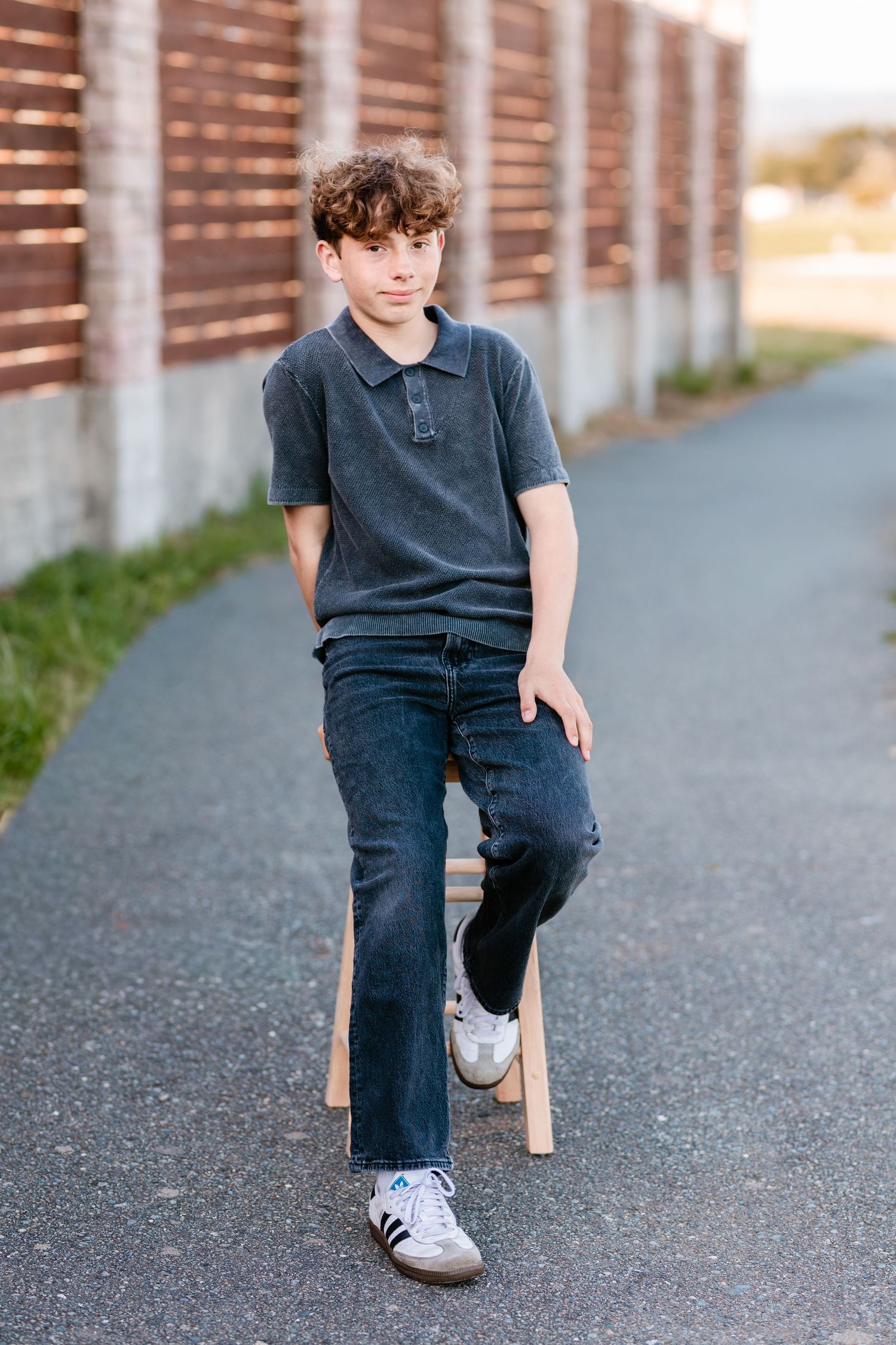 Teen boy in gray polo shirt and dark jeans sitting on wooden stool outdoors near brick fence.