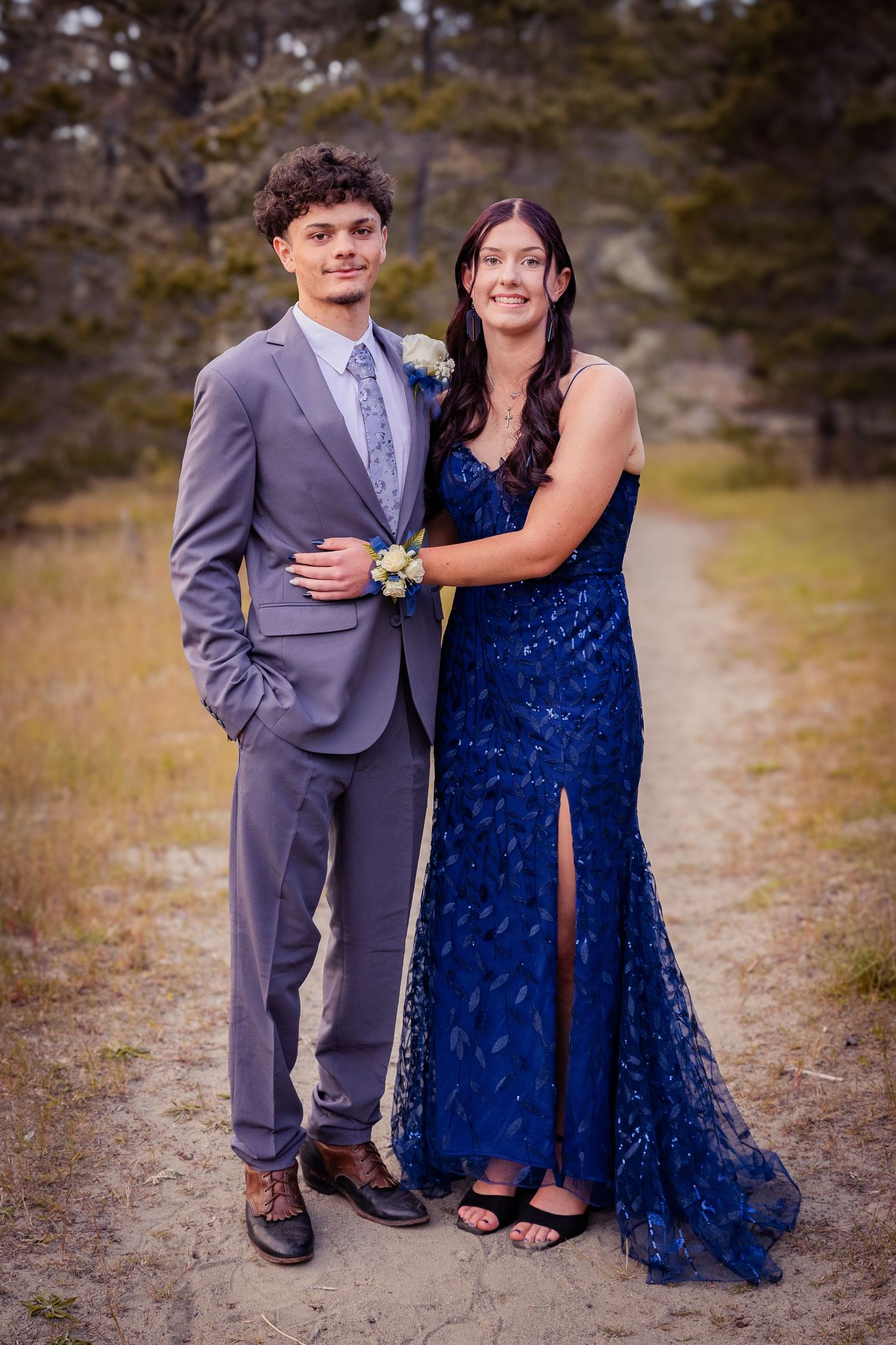 A couple poses for prom photos outdoors, wearing a blue sequin gown and gray suit with corsage.