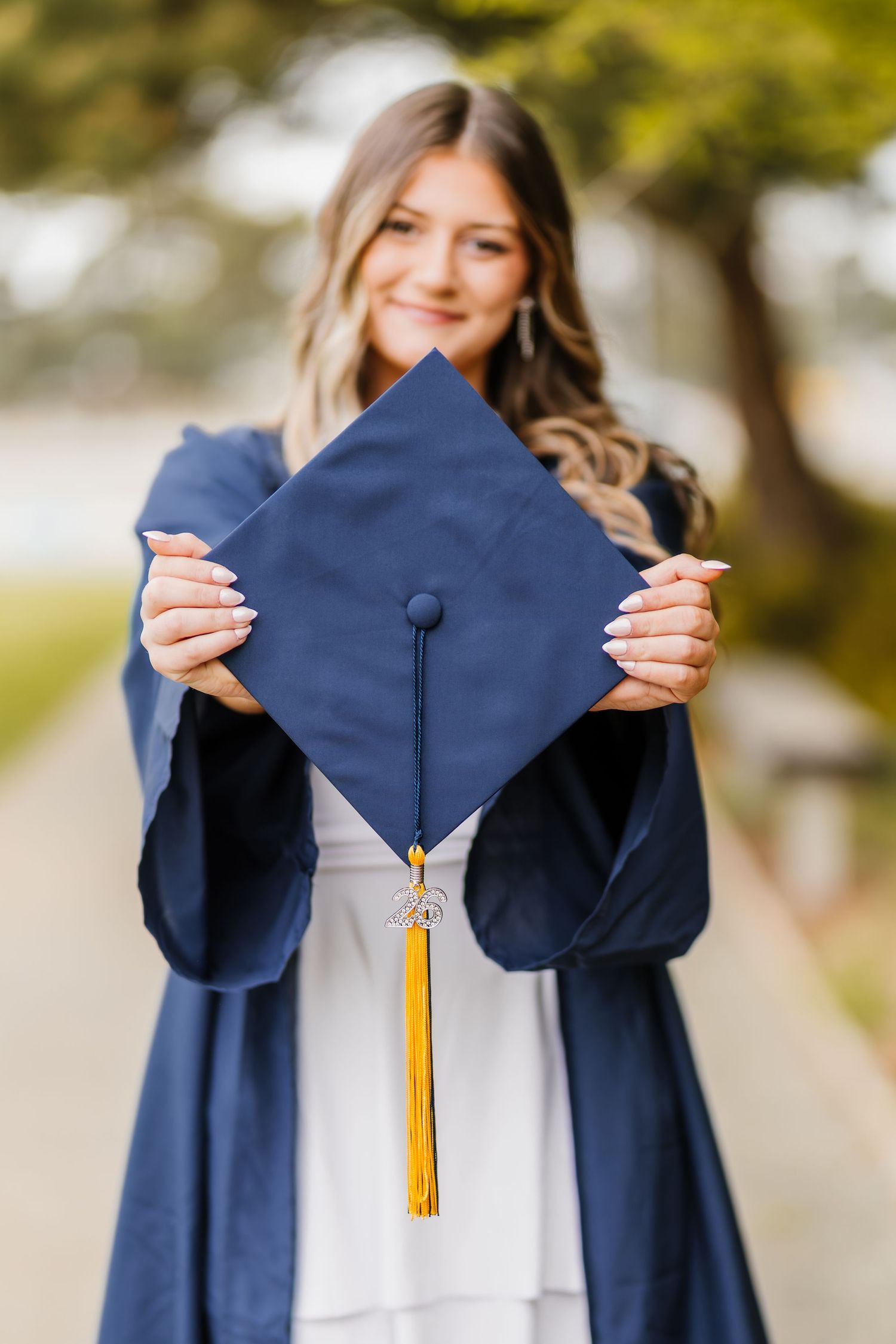 Young woman in navy graduation gown holding mortarboard cap with gold tassel outdoors.