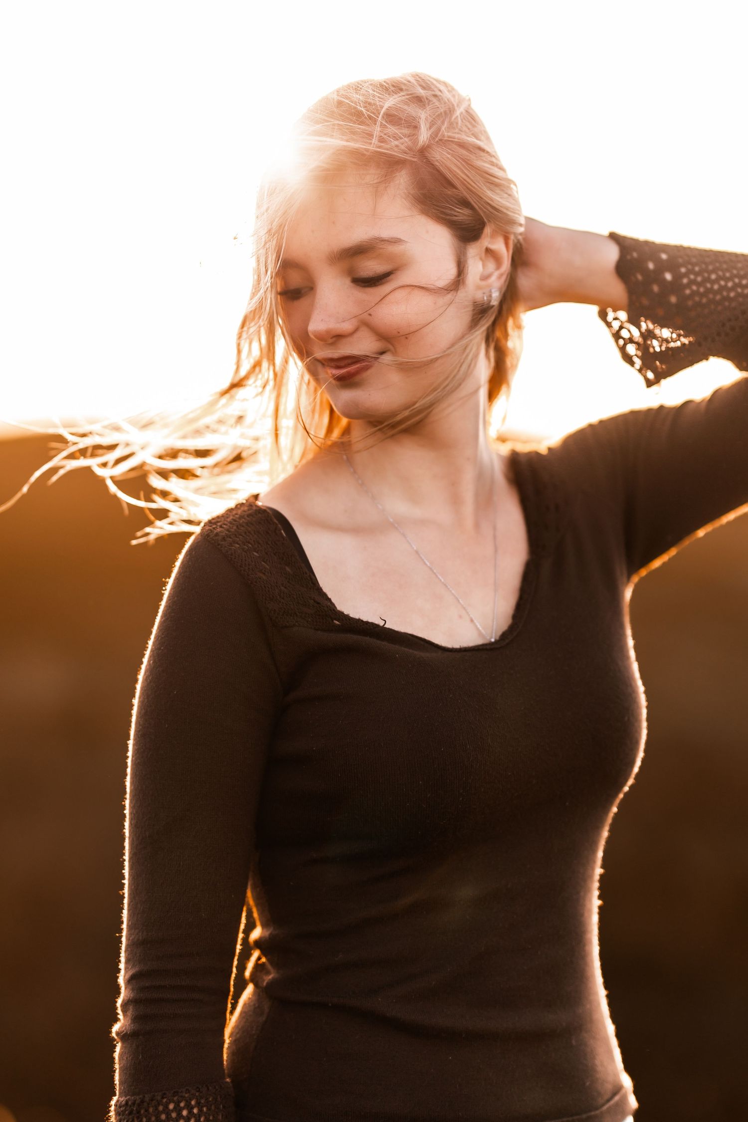 Blonde teen girl with eyes closed holding hair back in golden sunset light wearing black top.