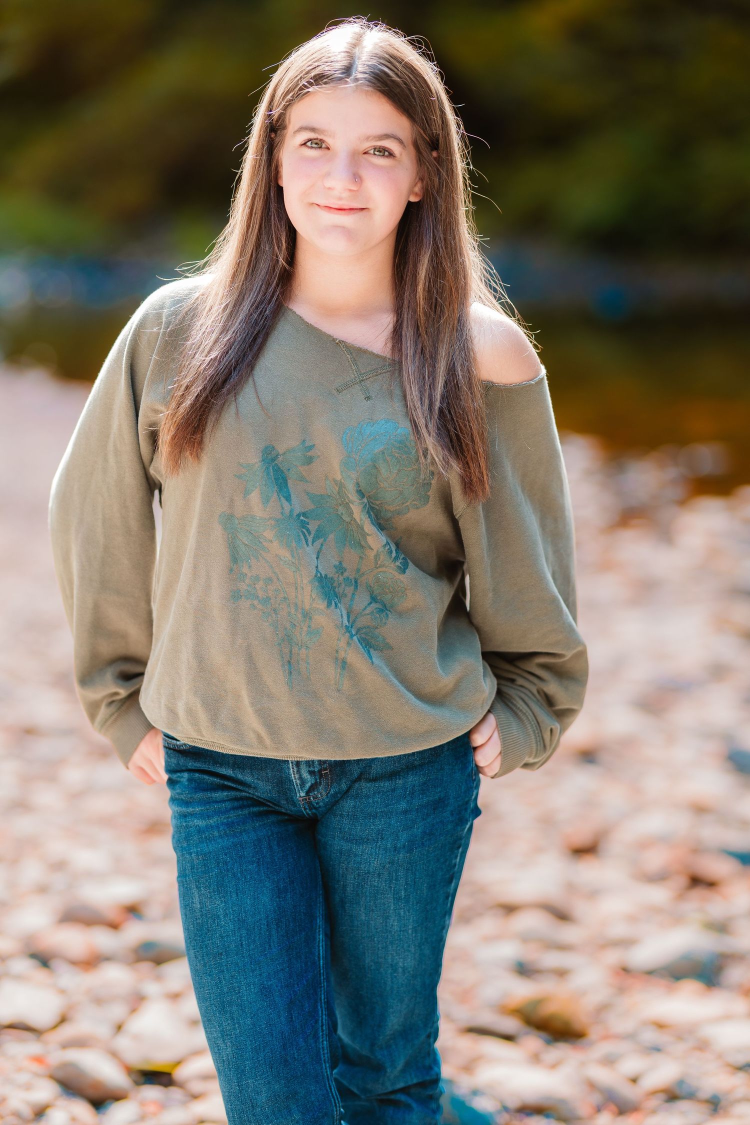 Teenage girl with long dark hair wearing a green sweatshirt and jeans, posing near a rocky riverbank.